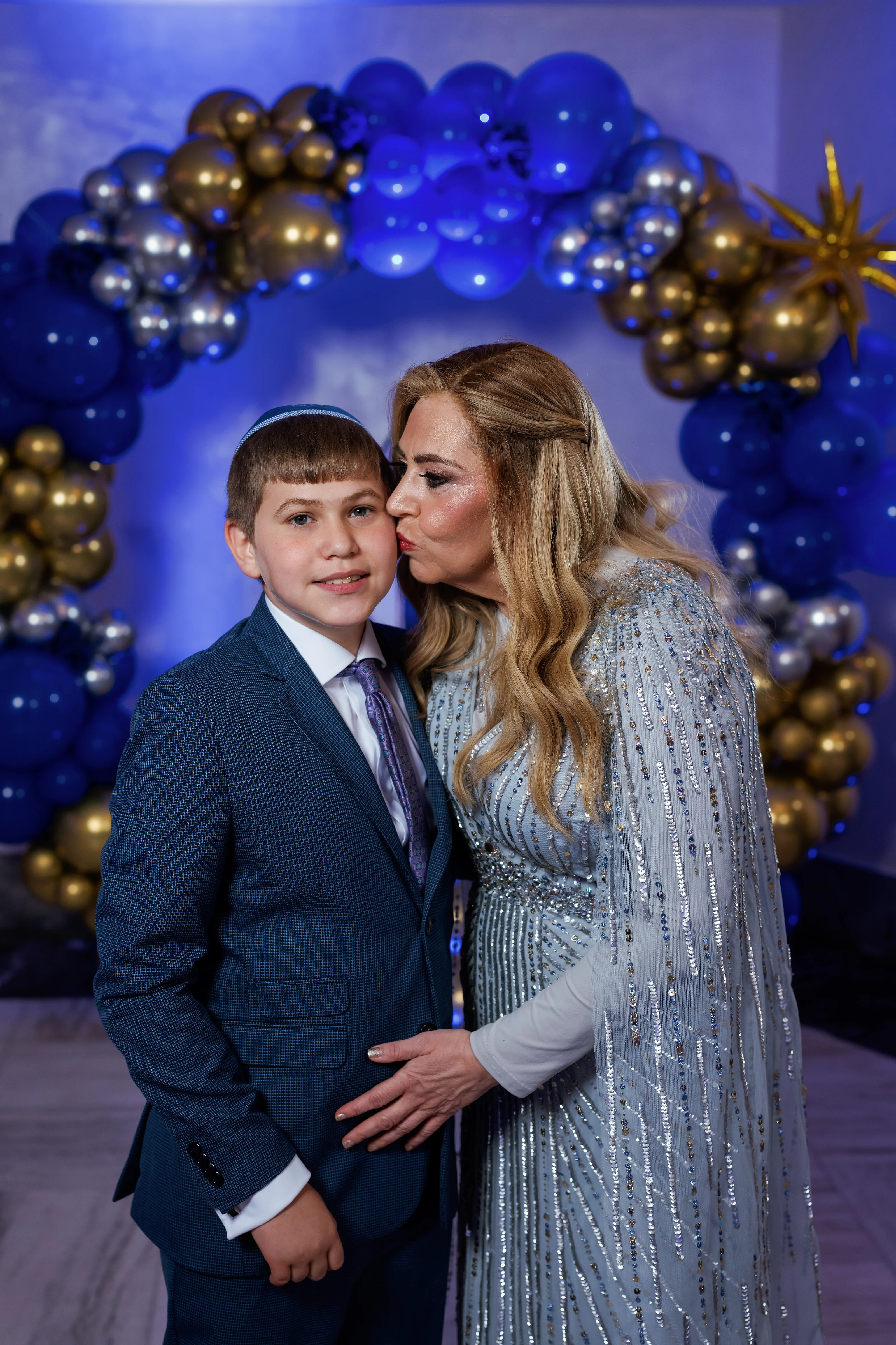 Portrait of a woman in a light sequined dress kissing a boy on the cheek in front of a blue and gold balloon backdrop.