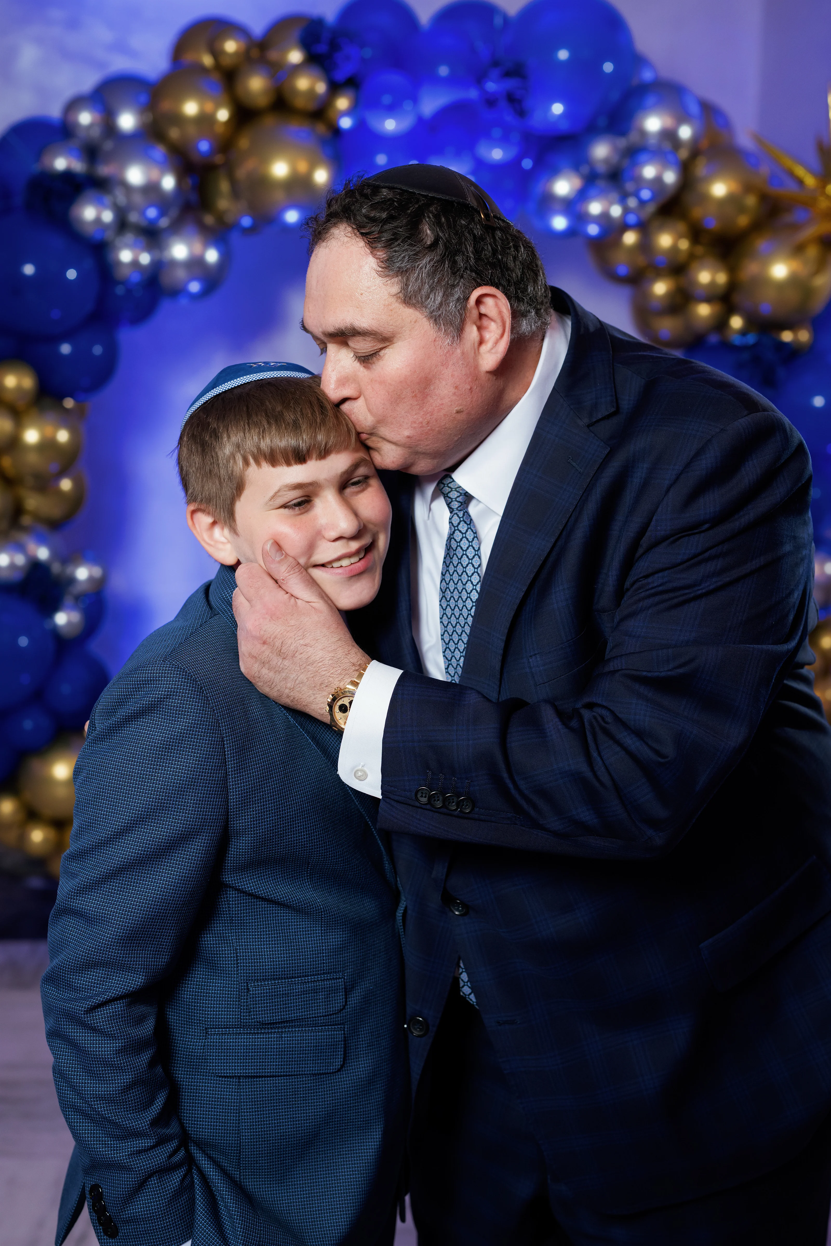 Close-up wedding moment of two people embracing with smiling expressions in front of blue and gold balloon décor.