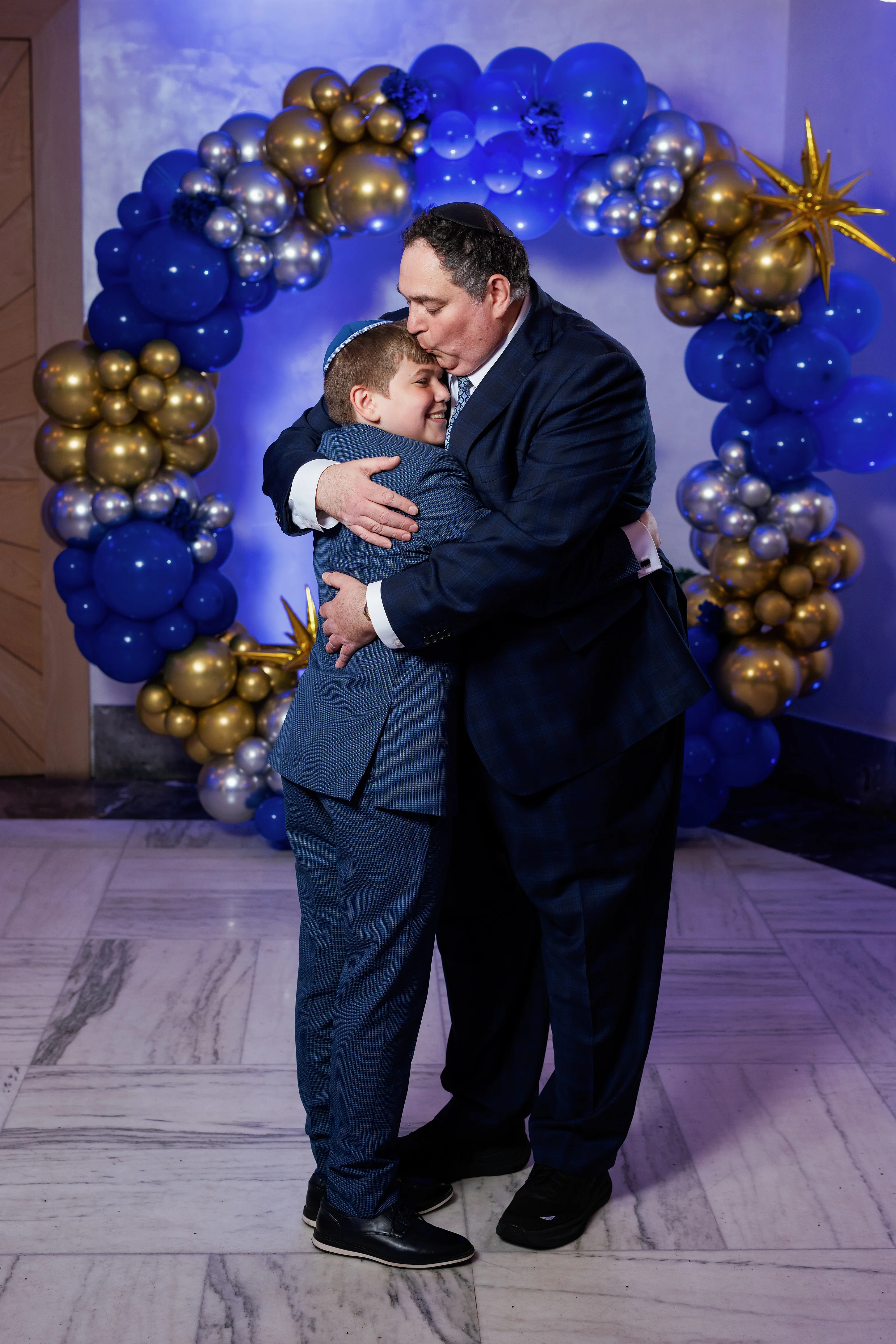 A man and a small child share an affectionate embrace in front of a blue and gold balloon backdrop during a celebration.