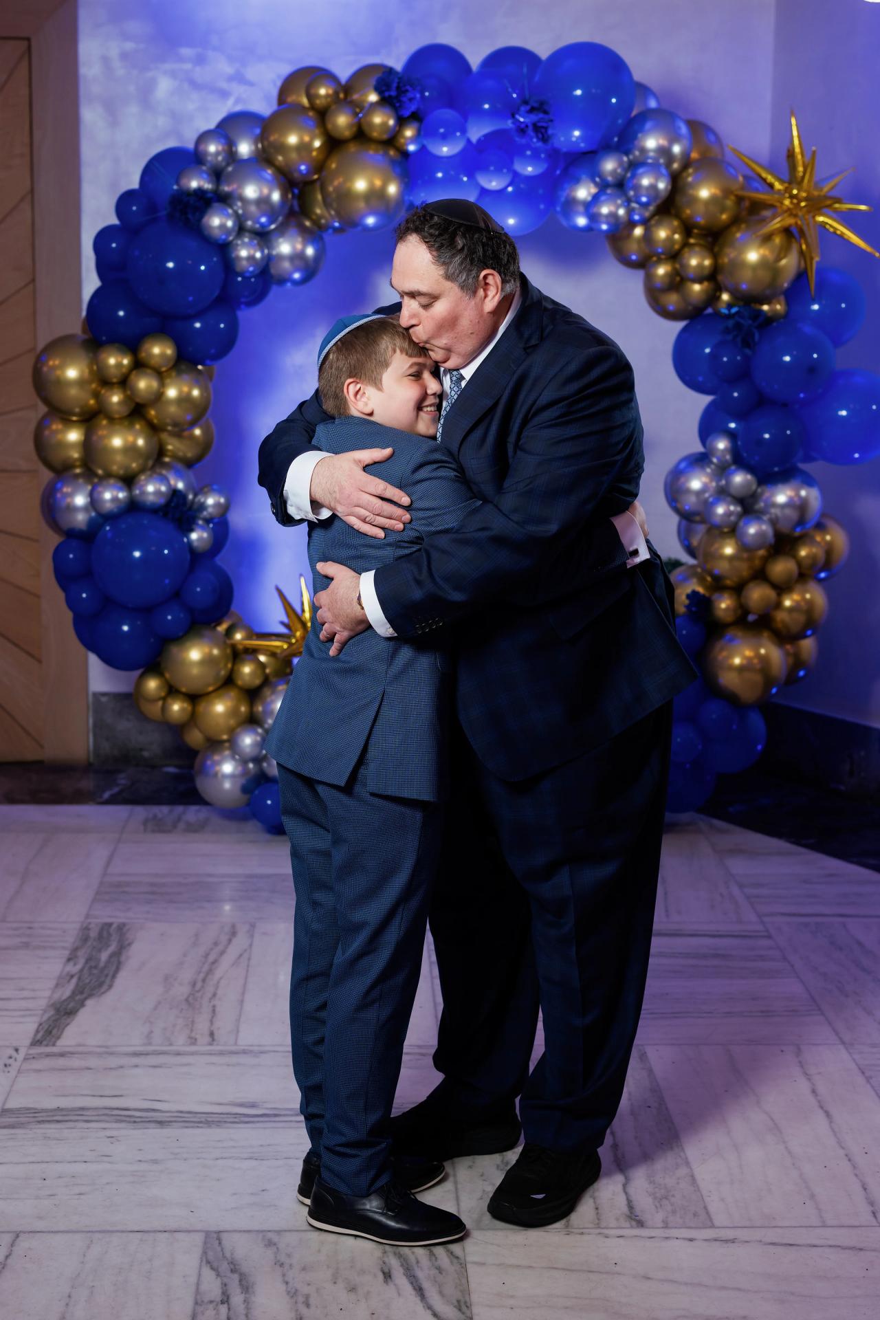 A man and a small child share an affectionate embrace in front of a blue and gold balloon backdrop during a celebration. Bar Mitzvah photography at Congregation Ohr Torah, Valley Stream, NY.