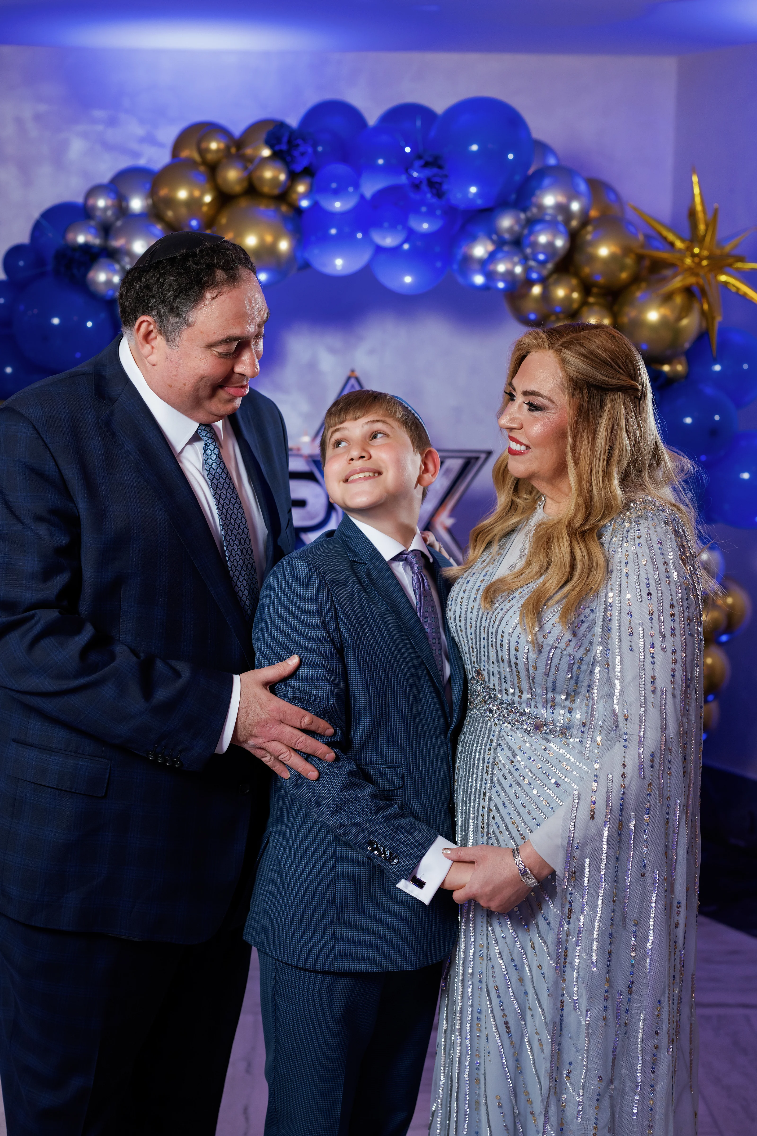 Bride and groom share a joyful close moment with a young boy at a celebration backdrop of blue and gold balloons.