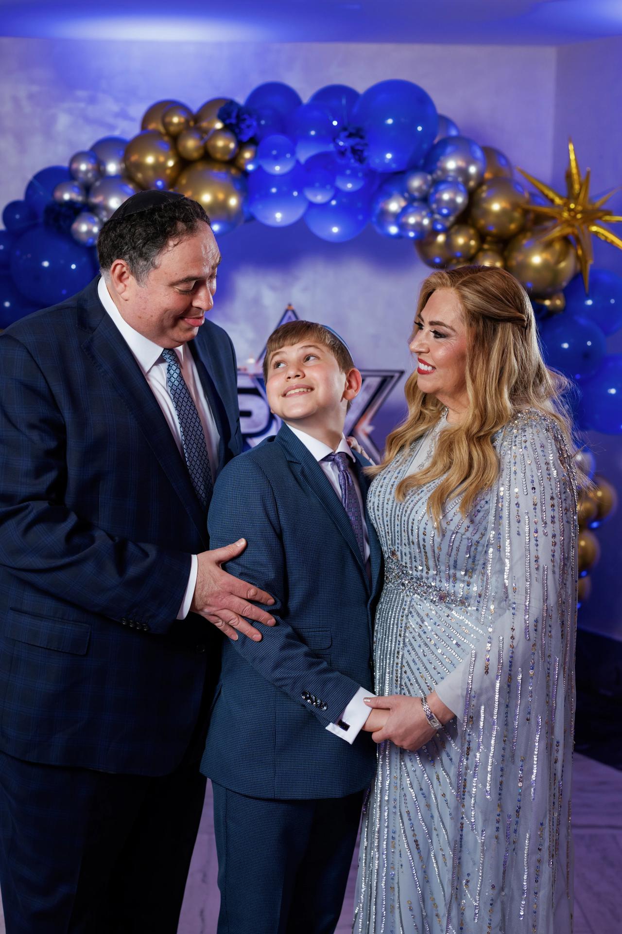 Bride and groom share a joyful close moment with a young boy at a celebration backdrop of blue and gold balloons. Bar Mitzvah photography at Congregation Ohr Torah, Valley Stream, NY.