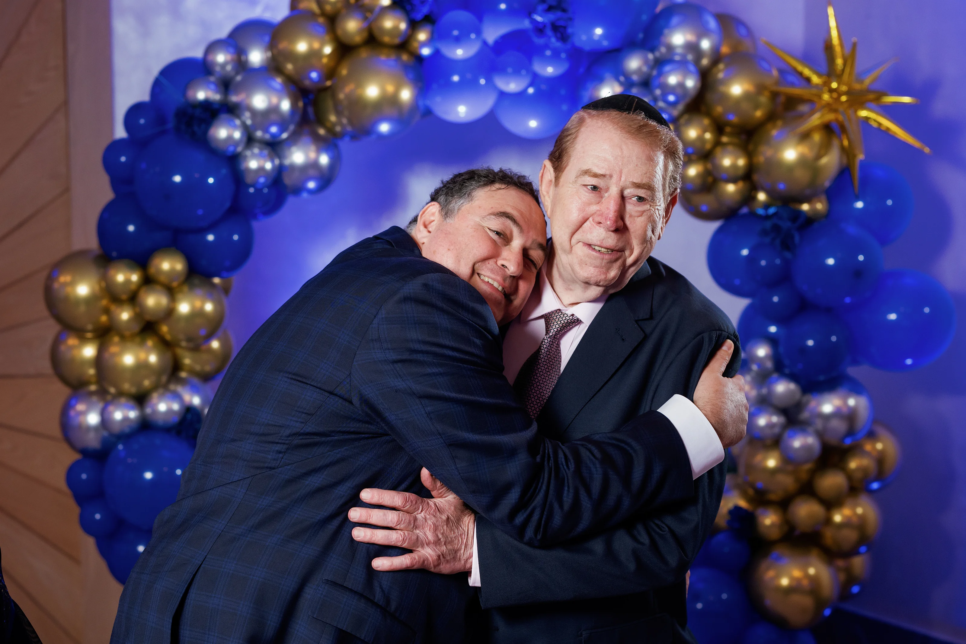 Two men in formal suits sharing an affectionate hug in front of a blue-and-gold balloon and star-themed backdrop.