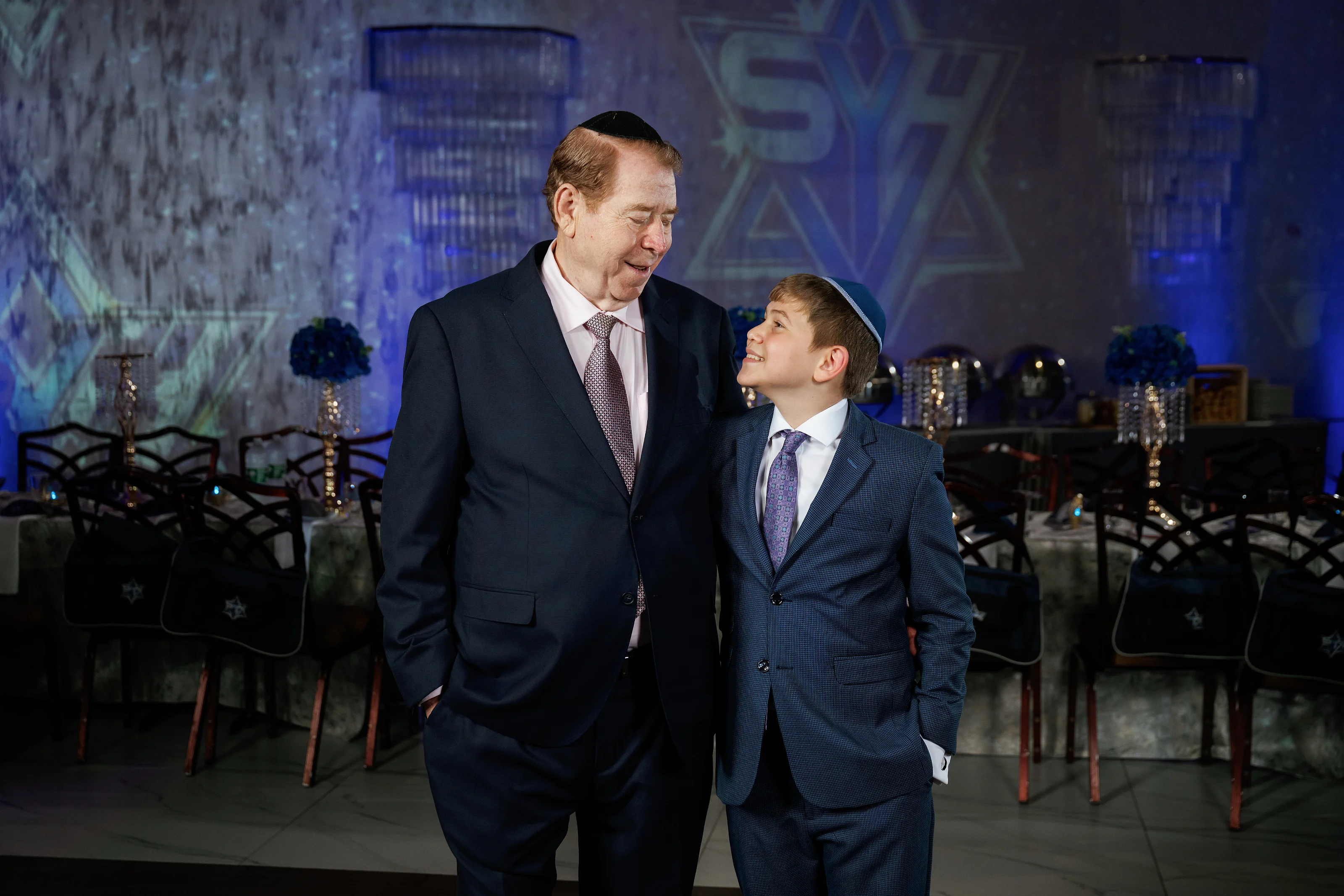 Man and young boy sharing a warm candid smile during a blue-lit wedding event, standing in front of decorated chairs and a lighted backdrop.