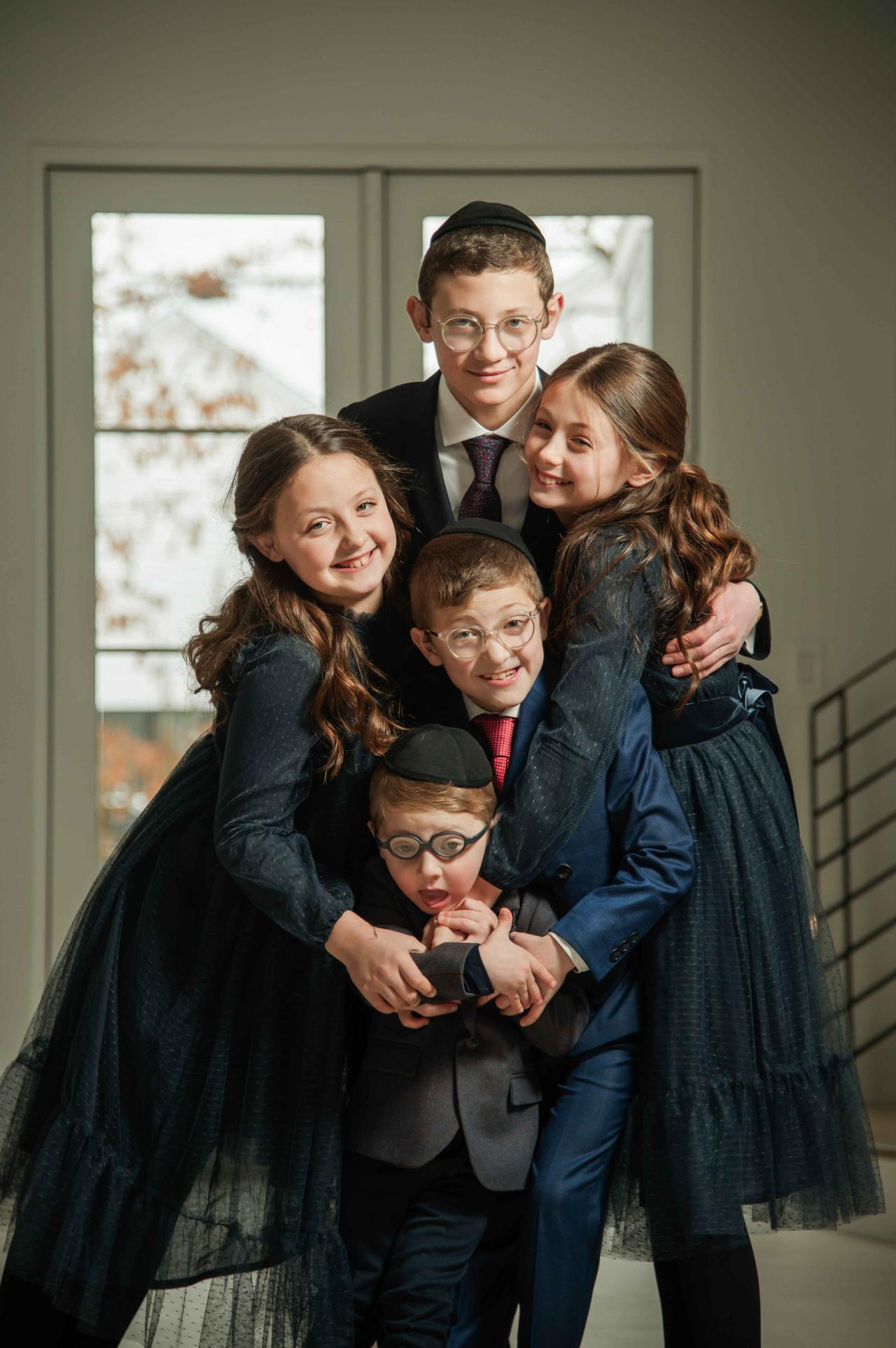Five children posing together in matching festive outfits, standing close with arms around each other indoors, smiling toward the camera.
