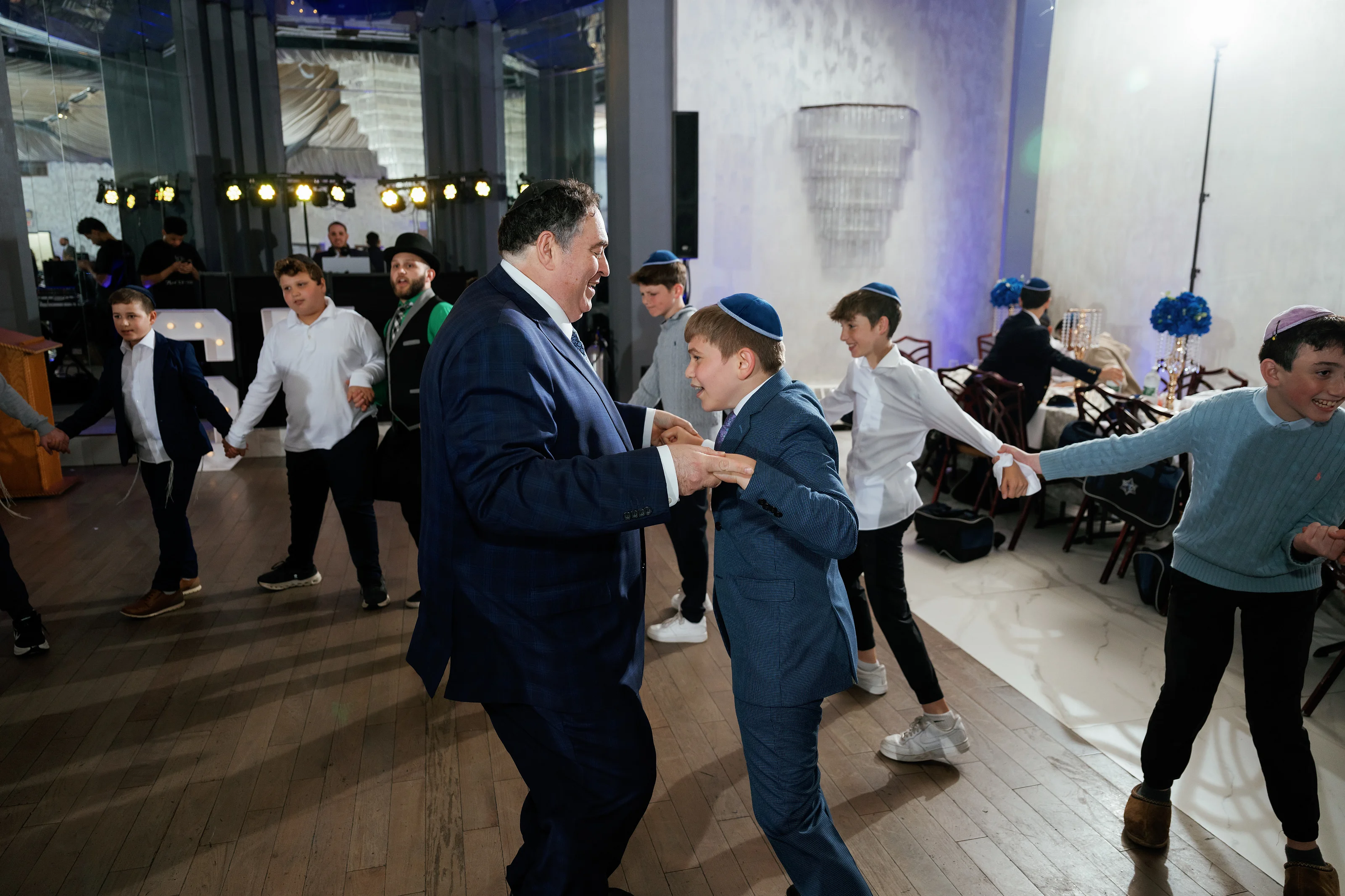 Groom dancing with a boy at a formal celebration with children and guests moving in the background under blue and warm lights.
