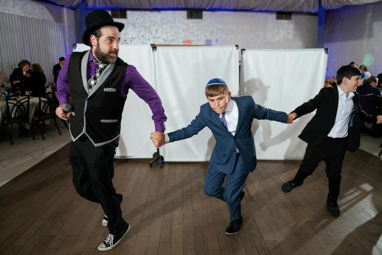 Candid dance-floor moment at a celebration with three men in formal attire moving in sync under blue accent lighting. Bar Mitzvah photography at Congregation Ohr Torah, Valley Stream, NY.