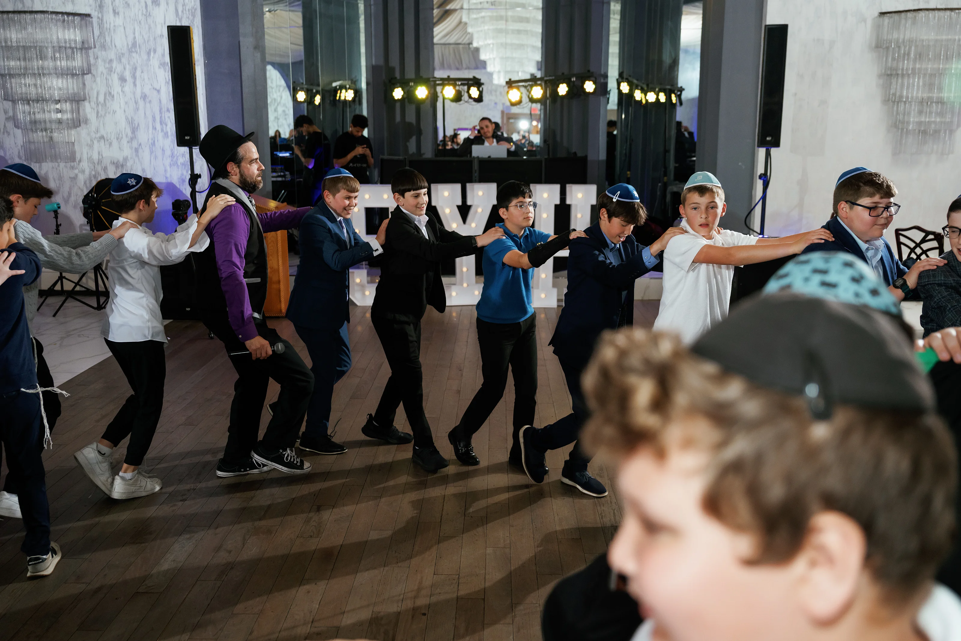 A group of guests dancing at a Jewish celebration, captured mid-motion in an indoor event hall with warm lighting.