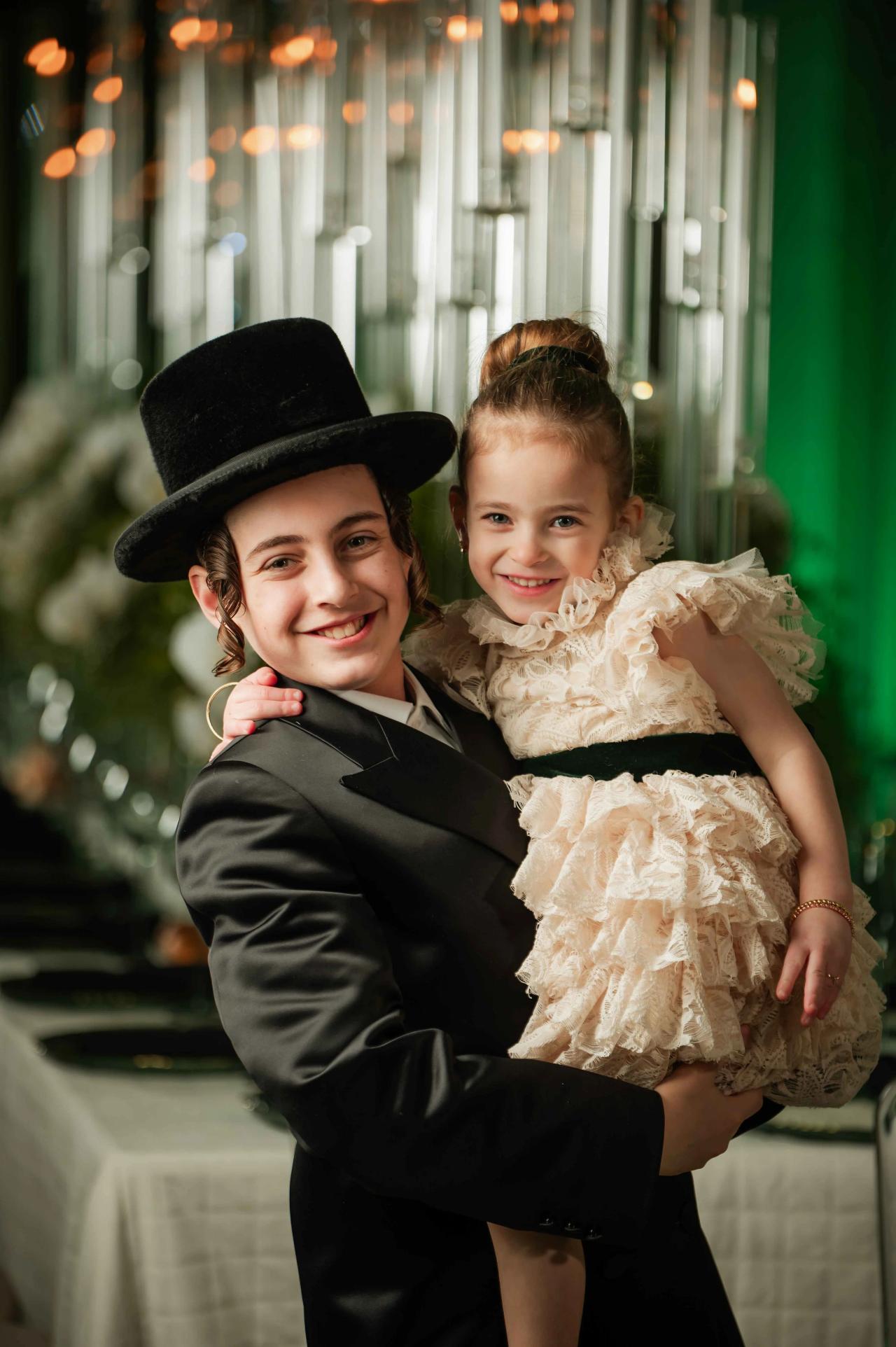 Smiling child in a black outfit and hat holds a younger girl in a cream ruffled dress during a warm, softly lit moment.