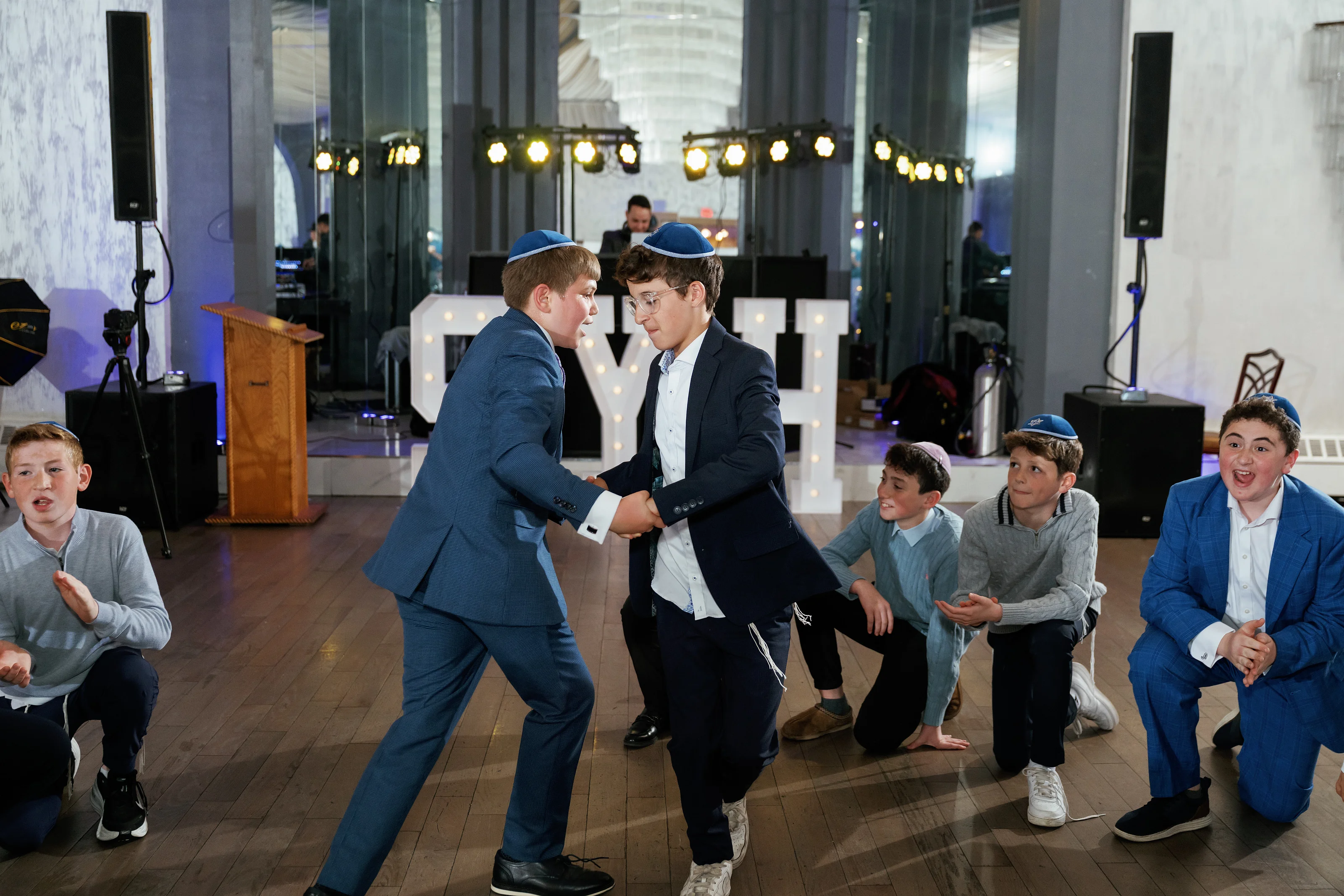 Two boys in kippot shaking hands and dancing while other children watch seated during a mitzvah celebration.