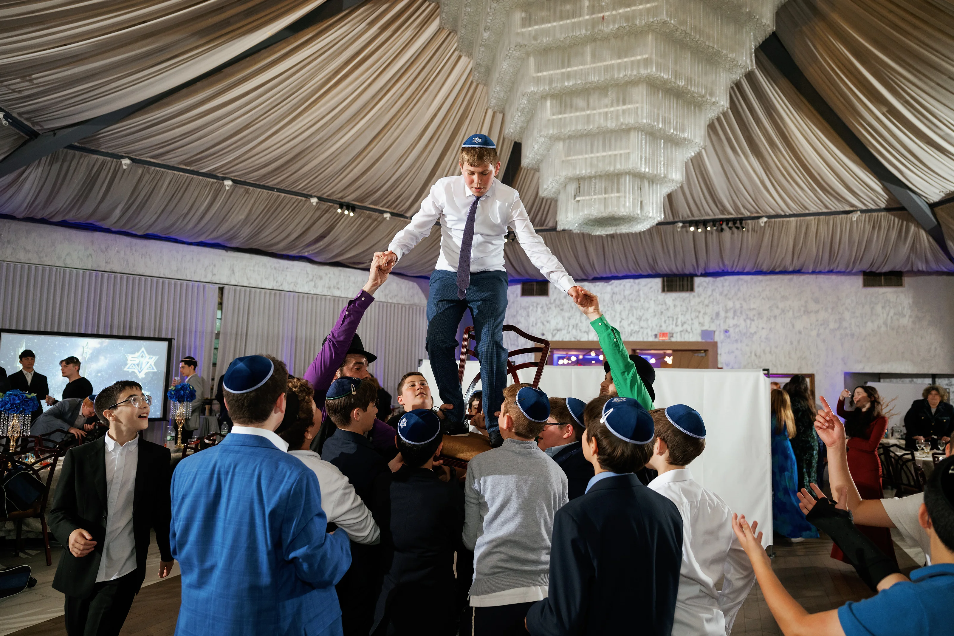 Guests dancing at an indoor celebration under a large canopy while a performer stands above the crowd with arms out.