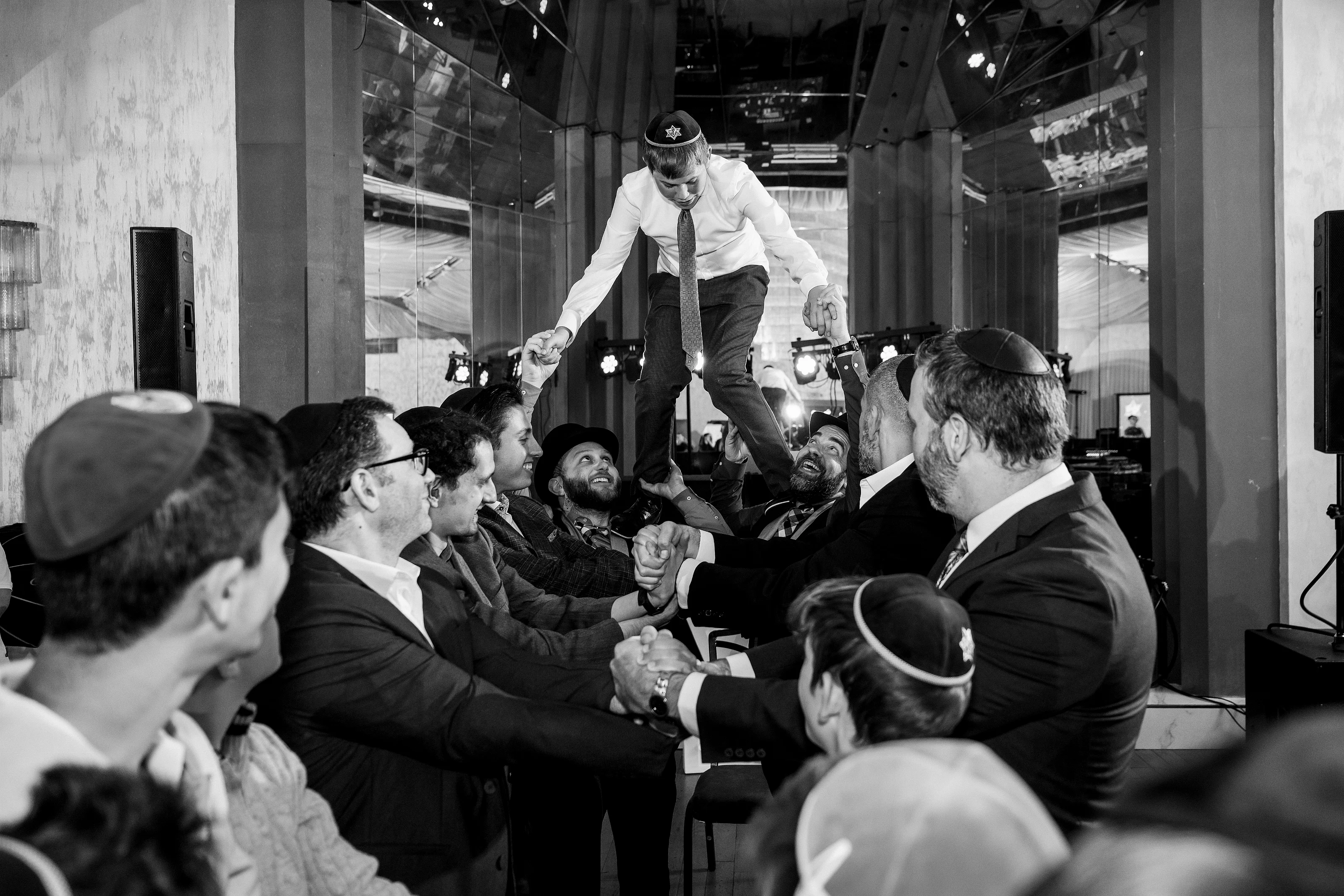 Black-and-white reception moment with a person lifted above a cheering crowd as guests reach and celebrate.