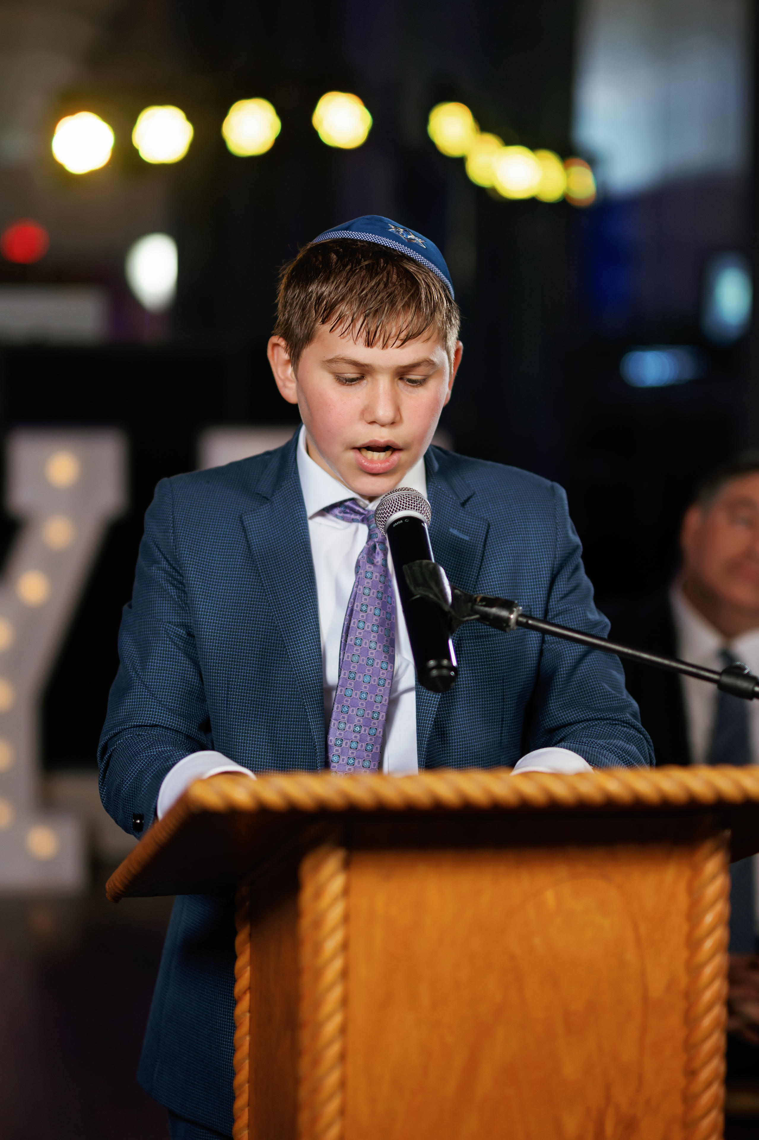 Young boy in a kippah and suit speaking into a microphone behind a wooden podium during a mitzvah moment.