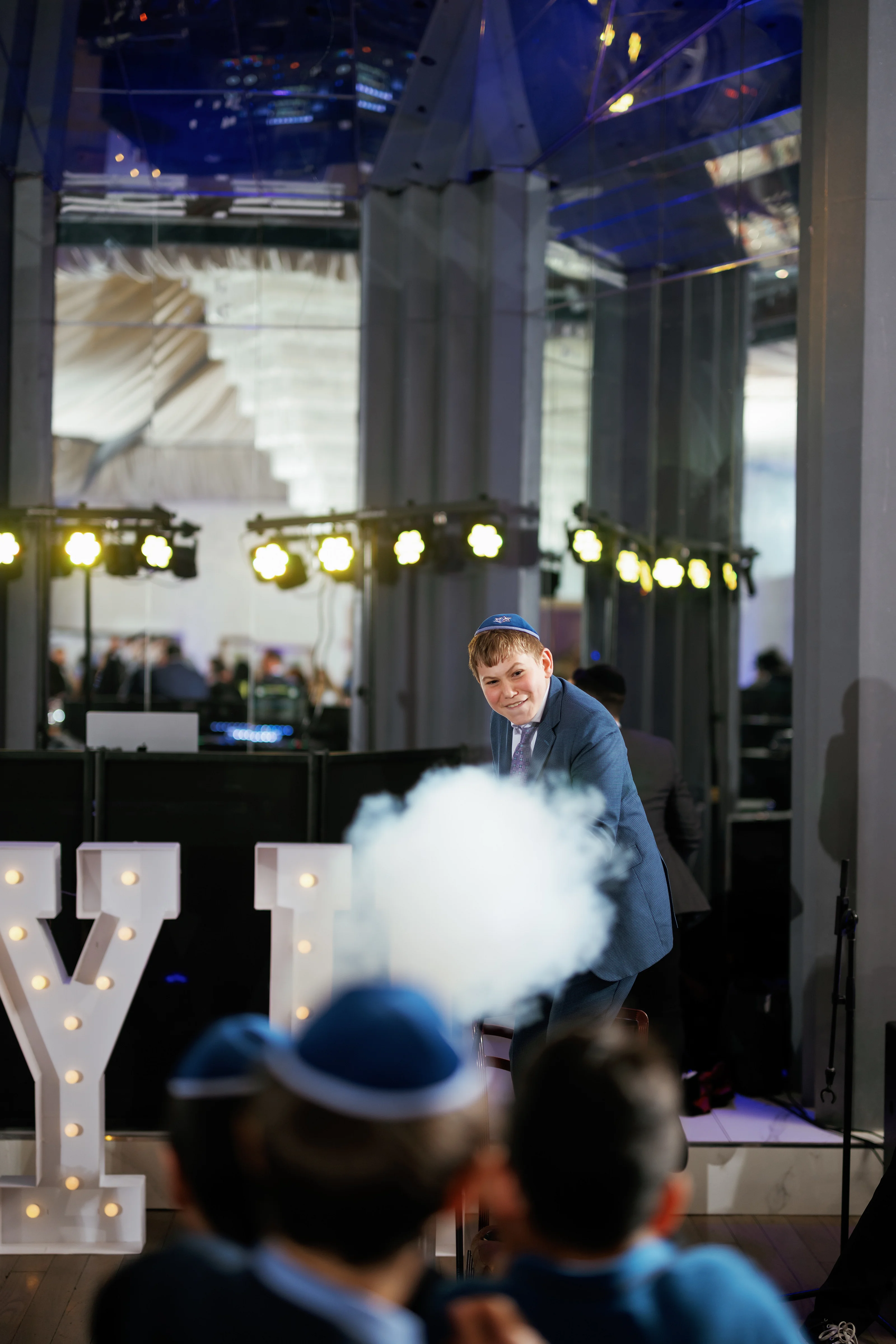 Smiling boy with a kippah leaning forward during a joyous wedding celebration, with stage lights, haze, and illuminated marquee letters in the background.