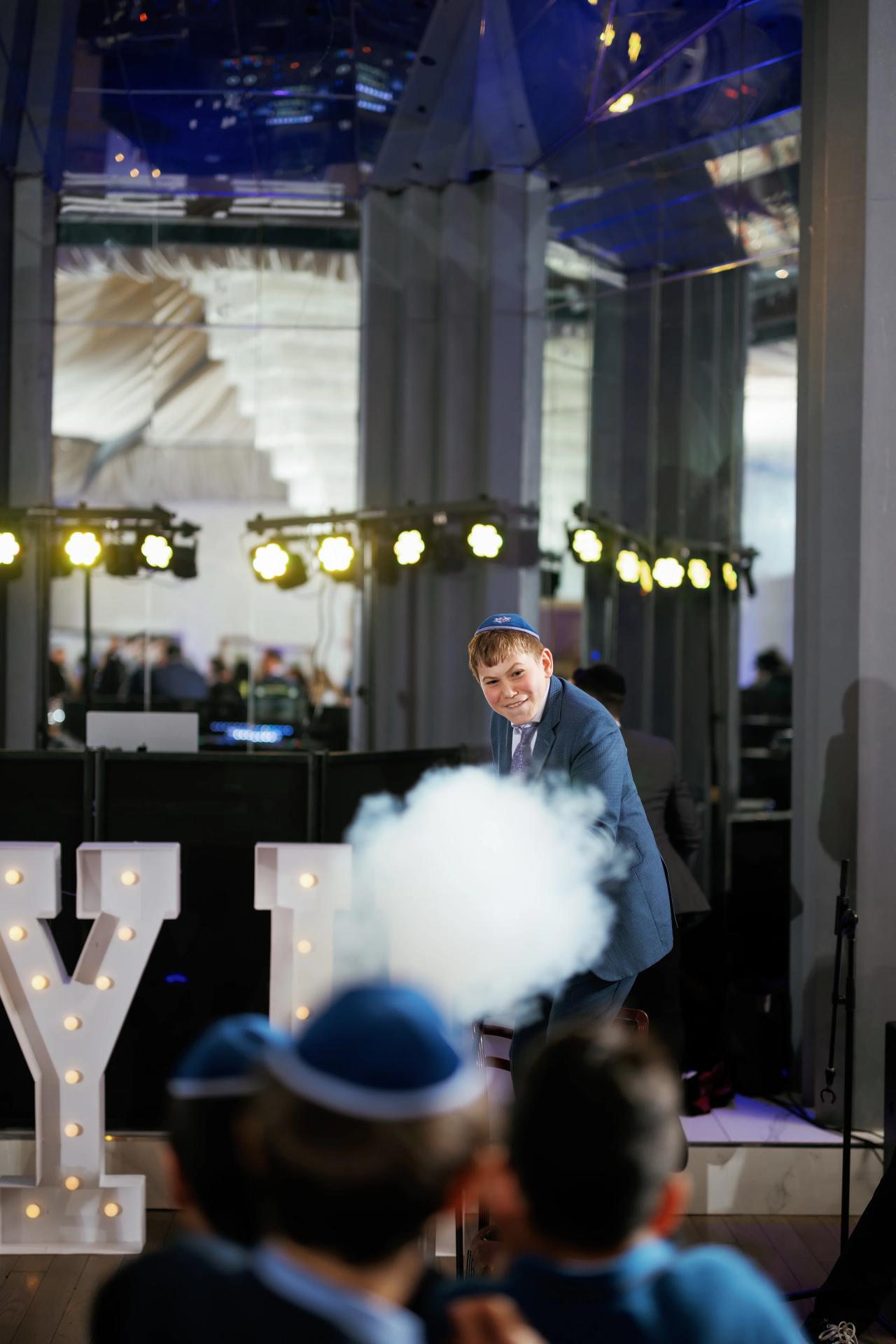 Smiling boy with a kippah leaning forward during a joyous wedding celebration, with stage lights, haze, and illuminated marquee letters in the background. Bar Mitzvah photography at Congregation Ohr Torah, Valley Stream, NY.