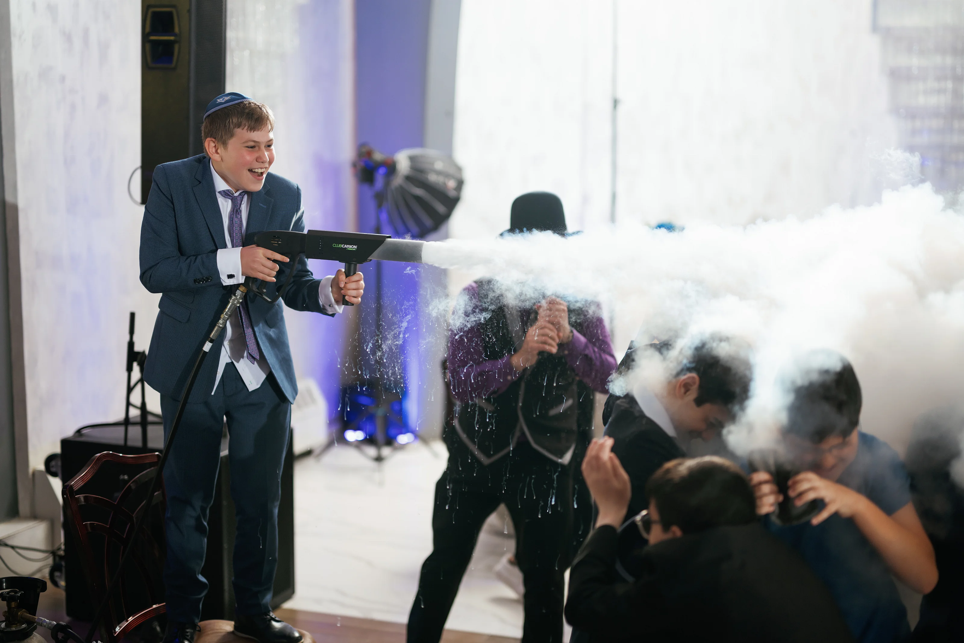 Smiling boy in a kippah in a suit at a wedding celebration while a confetti-like burst fills the air and adults react in the background.