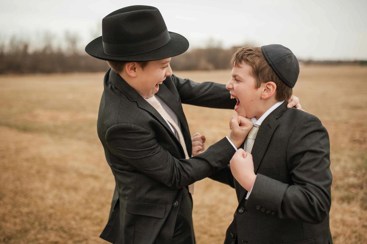 Outdoor candid of a man in a black hat smiling as he reaches to adjust a young boy’s collar and tefillin area while they stand in a field.