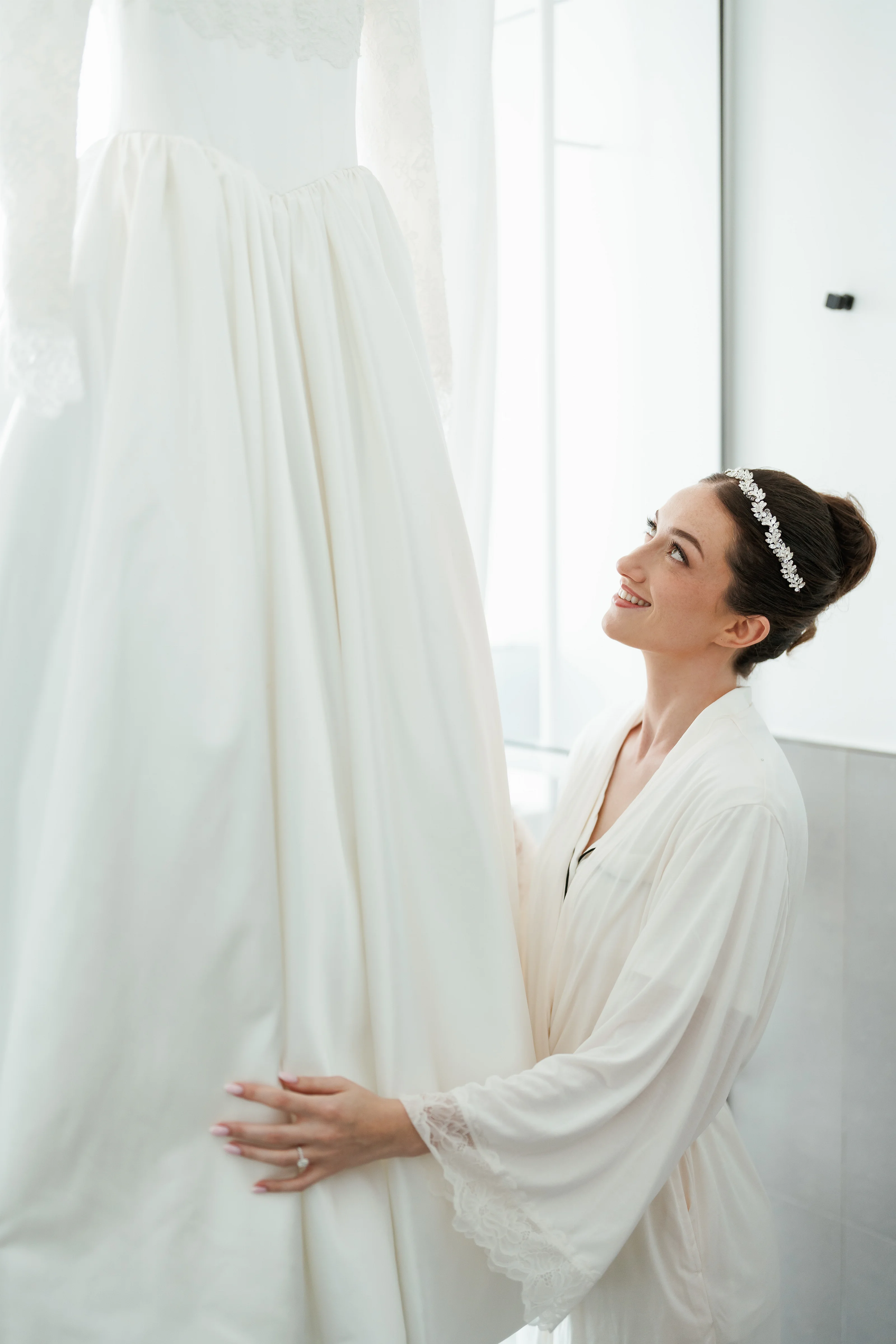 Bride in a white gown and jeweled headpiece smiling softly while holding her veil against a bright, minimalist background.