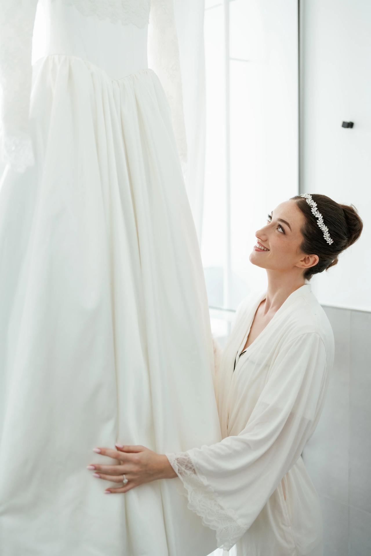 Bride in a white gown and jeweled headpiece smiling softly while holding her veil against a bright, minimalist background. Wedding at Ateres Chynka, Brooklyn, NY.