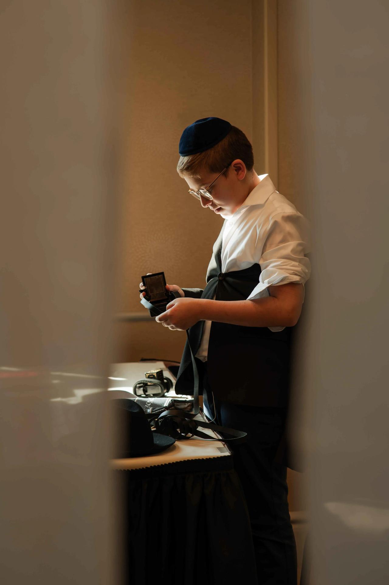 A boy in a kippah and white shirt stands holding and checking a tefillin box, looking down as light falls across the scene.