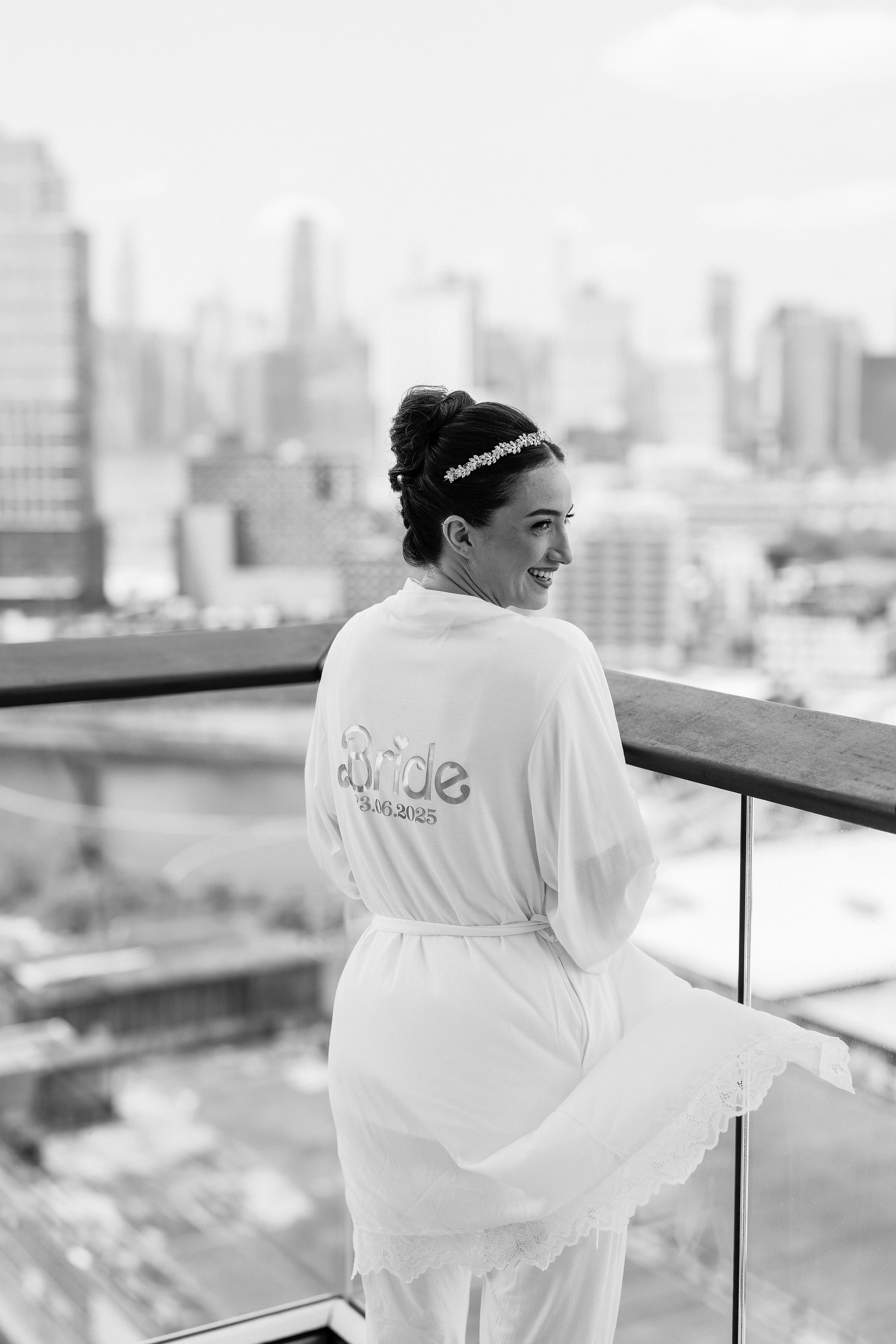 Black-and-white portrait of a bride in profile standing by a balcony railing with a city skyline in the background.