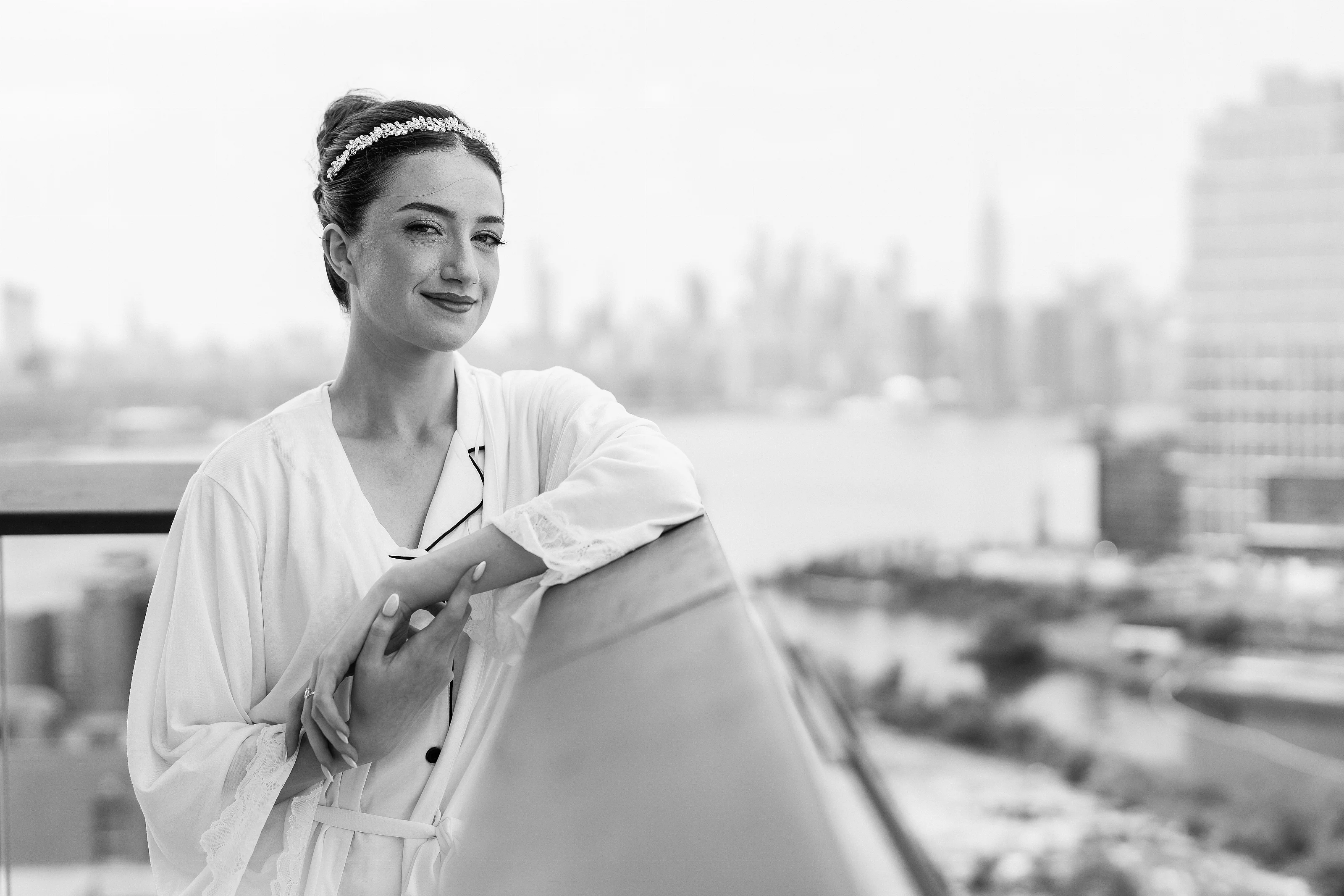 Black-and-white portrait of a bride wearing a white robe and sparkling headband, hands over the heart with a soft blurred city backdrop.