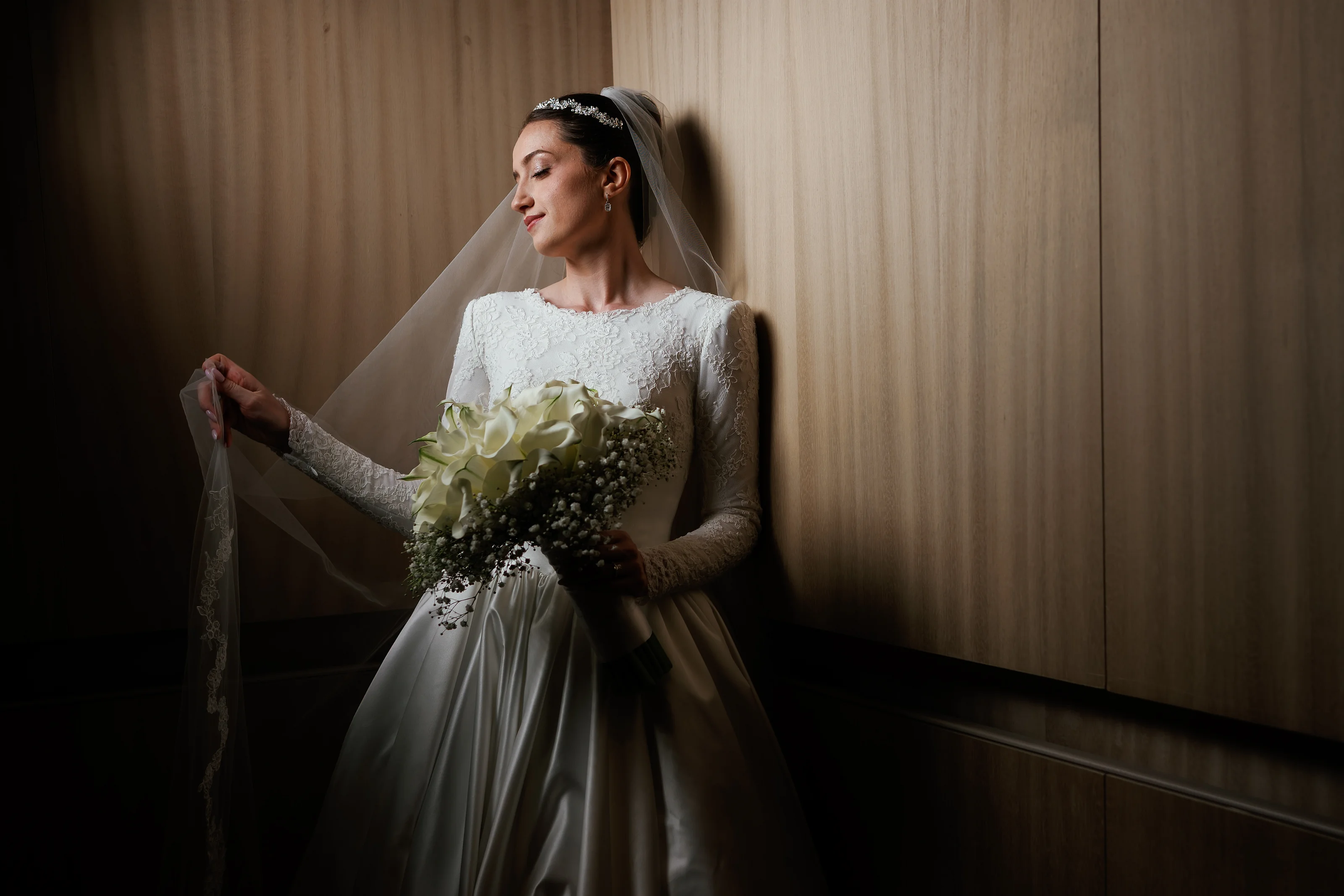 Bride in white lace dress holding her veil and bouquet in warm indoor light against wood paneling.
