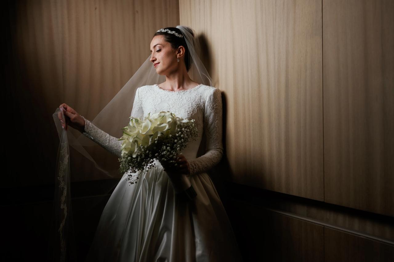 Bride in white lace dress holding her veil and bouquet in warm indoor light against wood paneling. Wedding at Ateres Chynka, Brooklyn, NY.