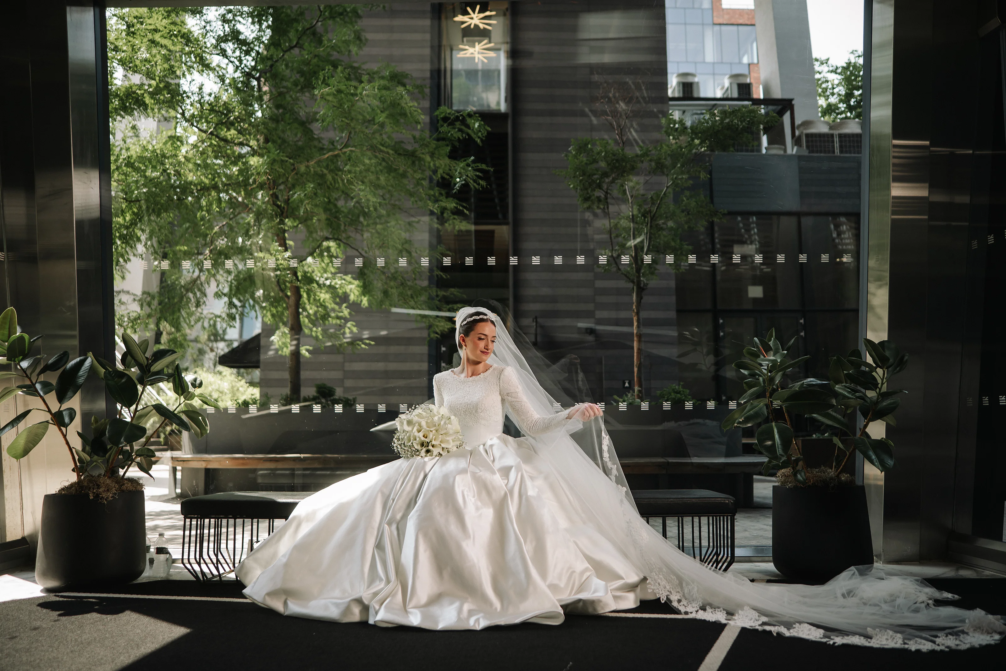Bride seated indoors in strong sunlight, wearing an ornate white wedding gown with a veil, greenery and modern architecture in the background.