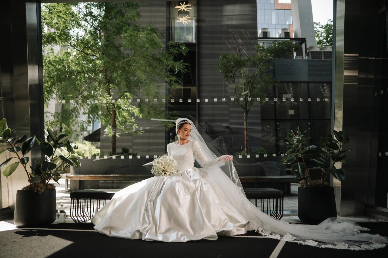 Bride seated indoors in strong sunlight, wearing an ornate white wedding gown with a veil, greenery and modern architecture in the background. Wedding at Ateres Chynka, Brooklyn, NY.