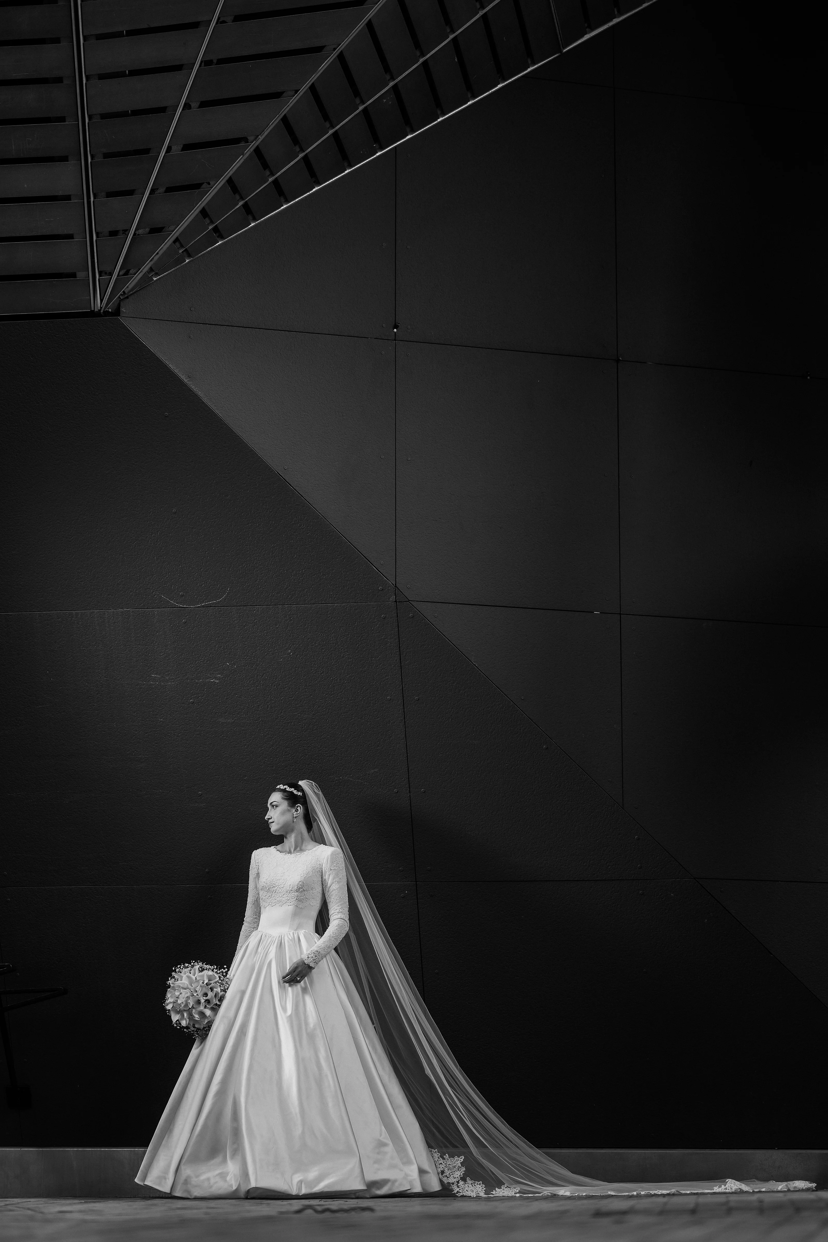Black-and-white editorial-style portrait of a bride in a bridal gown with a long veil flowing behind her against a dark geometric wall.