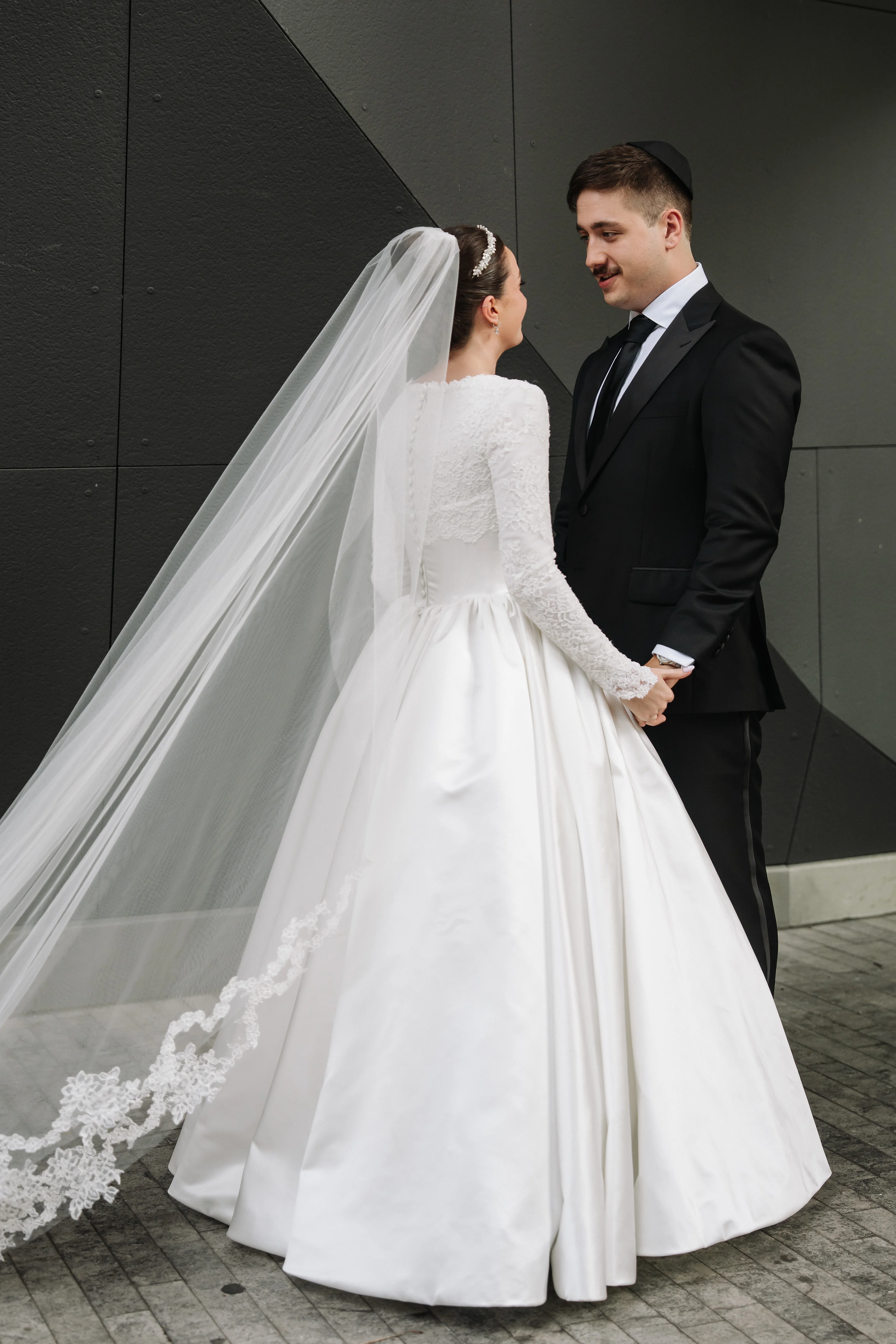 Bride and groom in wedding attire holding hands and smiling as the bride’s veil flows against a dark architectural wall background.