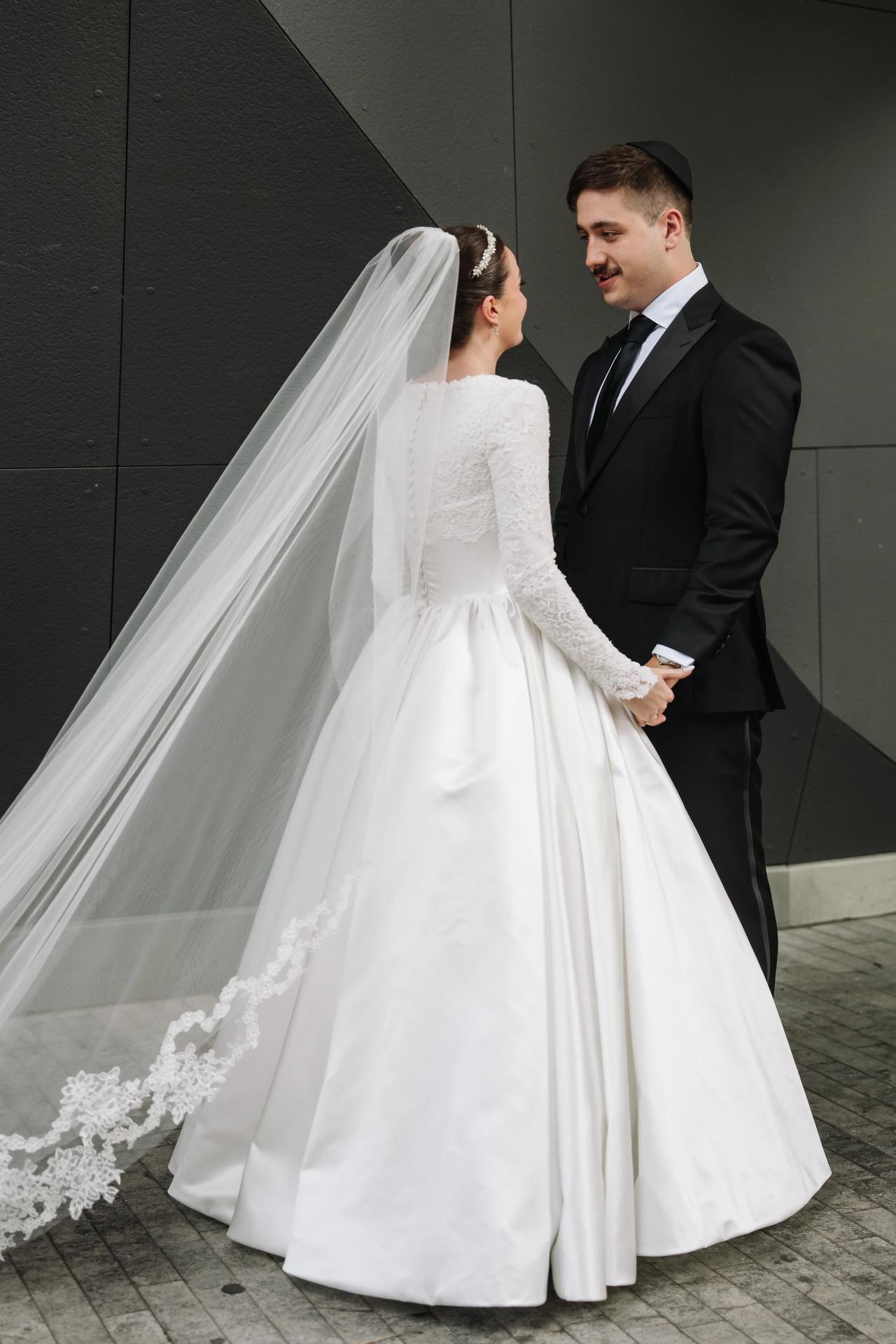 Bride and groom in wedding attire holding hands and smiling as the bride’s veil flows against a dark architectural wall background. Wedding at Ateres Chynka, Brooklyn, NY.