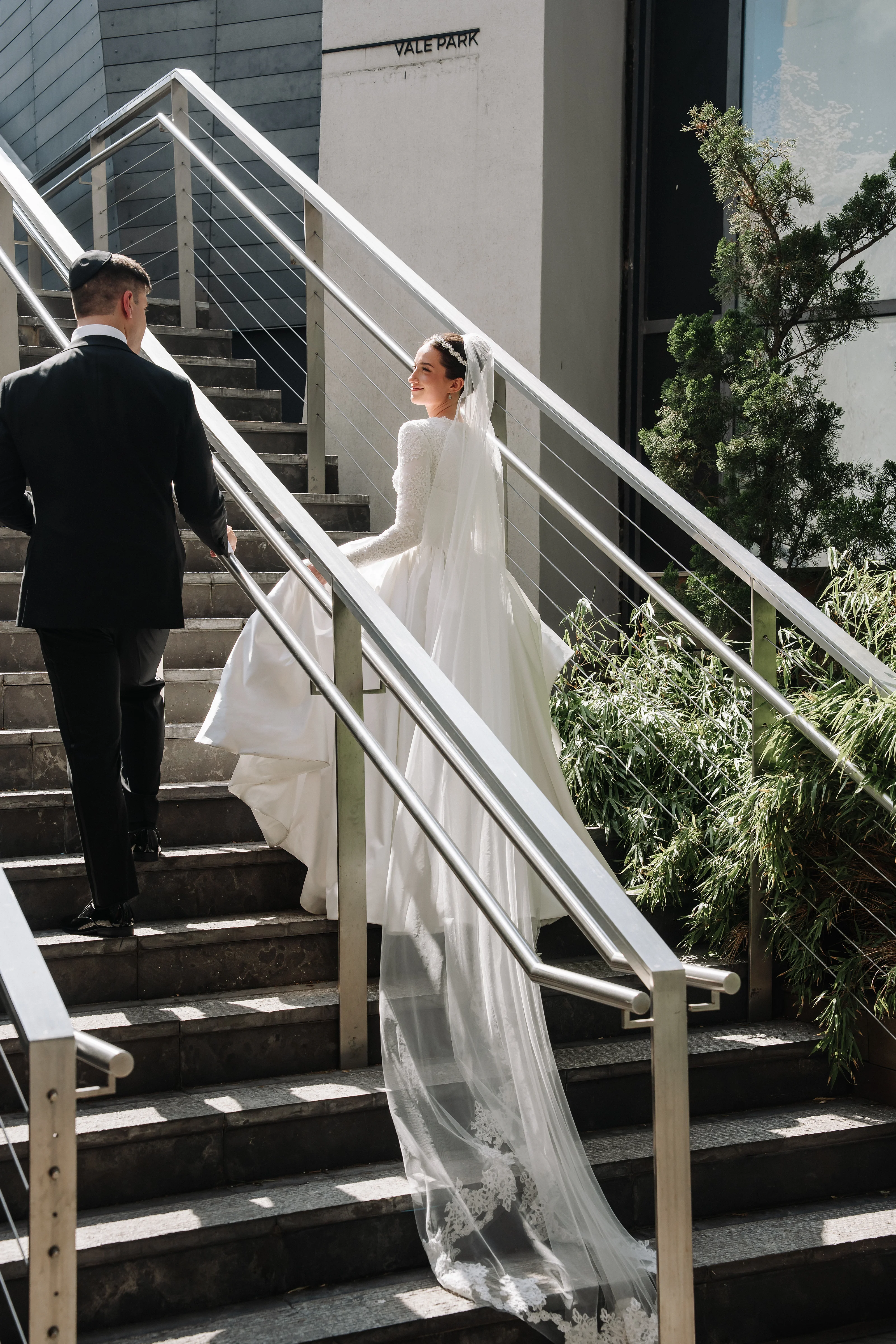 Bride descending outdoor stone steps in a flowing veil and lace gown, holding hands with groom beside her in bright daylight.
