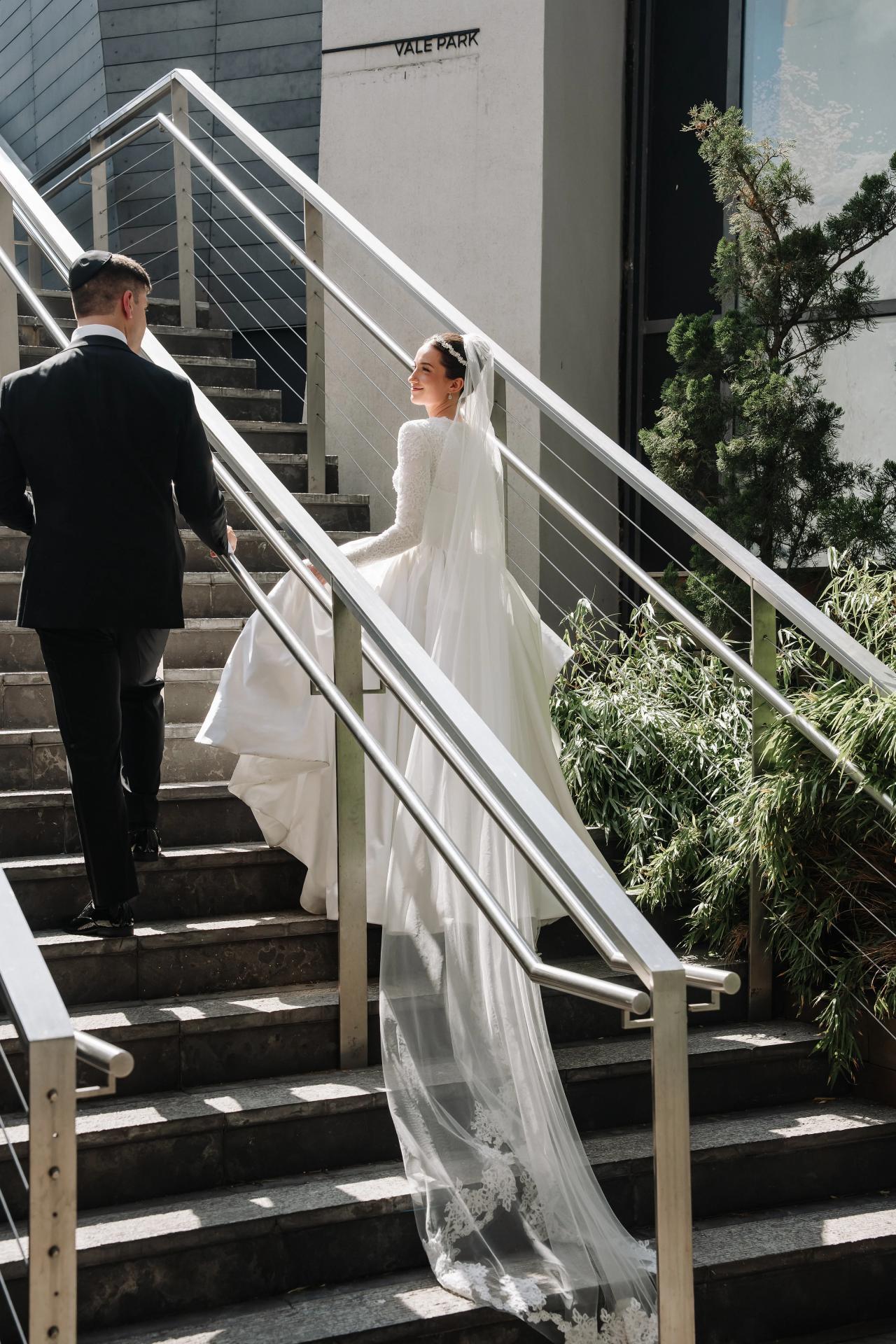 Bride descending outdoor stone steps in a flowing veil and lace gown, holding hands with groom beside her in bright daylight. Wedding at Ateres Chynka, Brooklyn, NY.