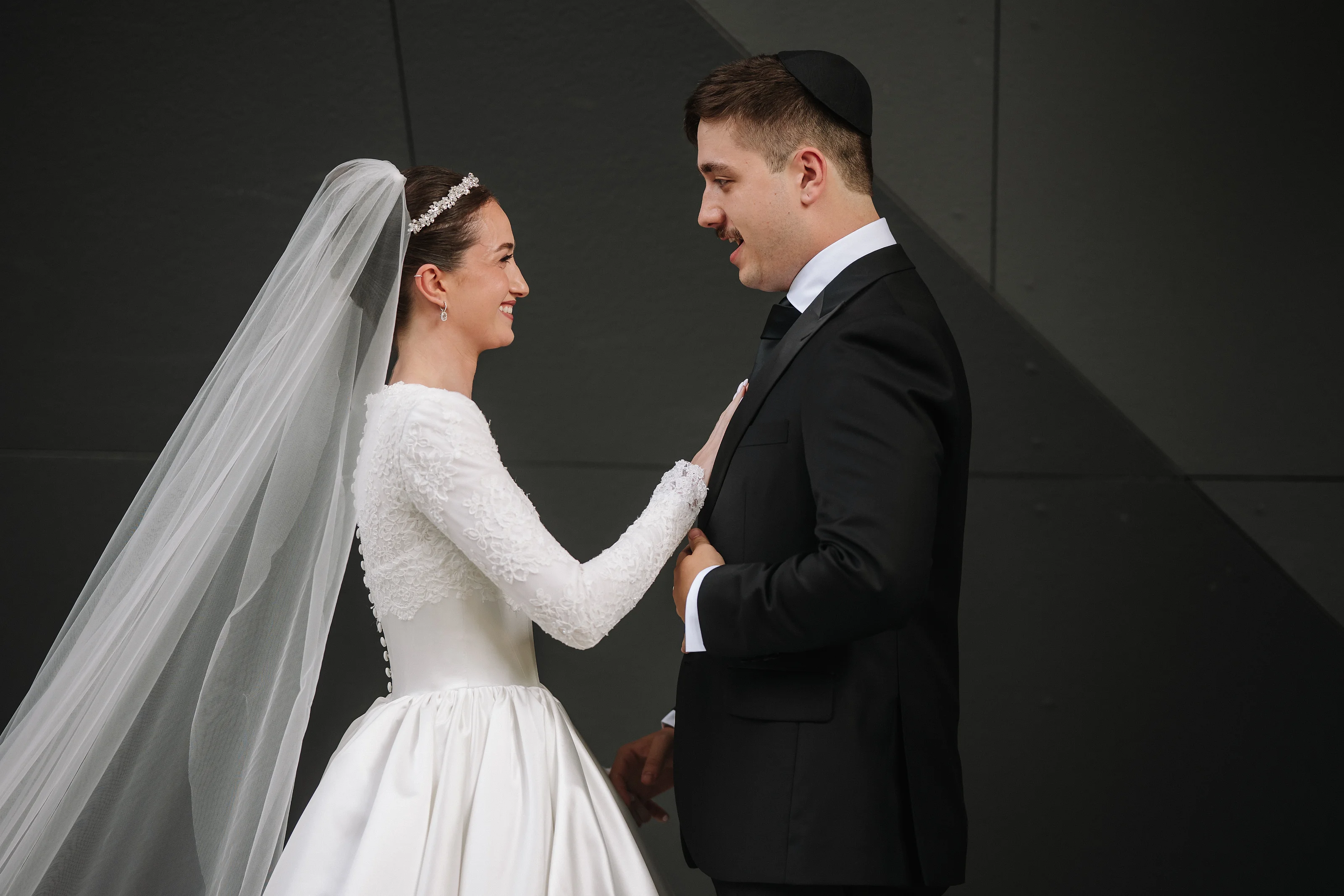 Bride in white lace gown and veil reaching for the groom’s hand in a quiet embrace against a dark gray wall.
