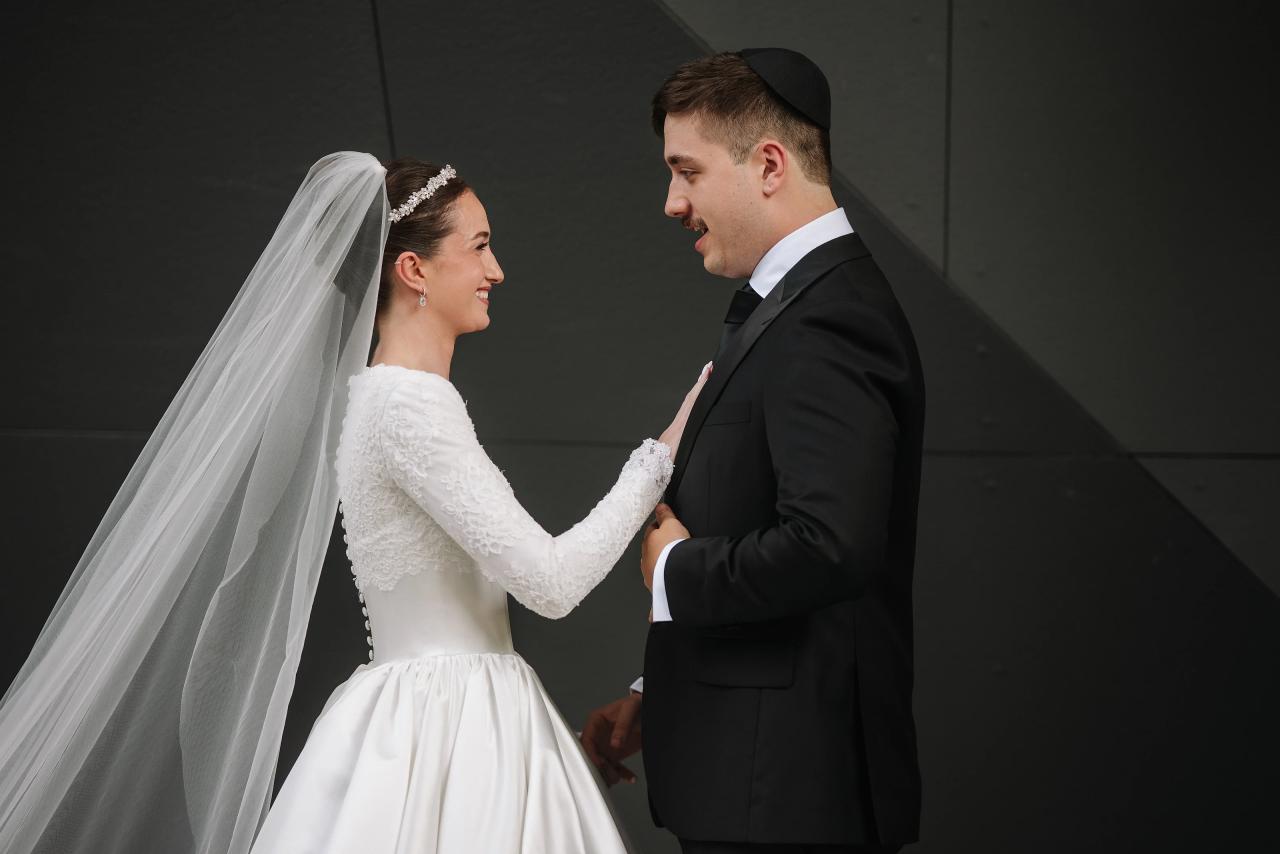 Bride in white lace gown and veil reaching for the groom’s hand in a quiet embrace against a dark gray wall. Wedding at Ateres Chynka, Brooklyn, NY.
