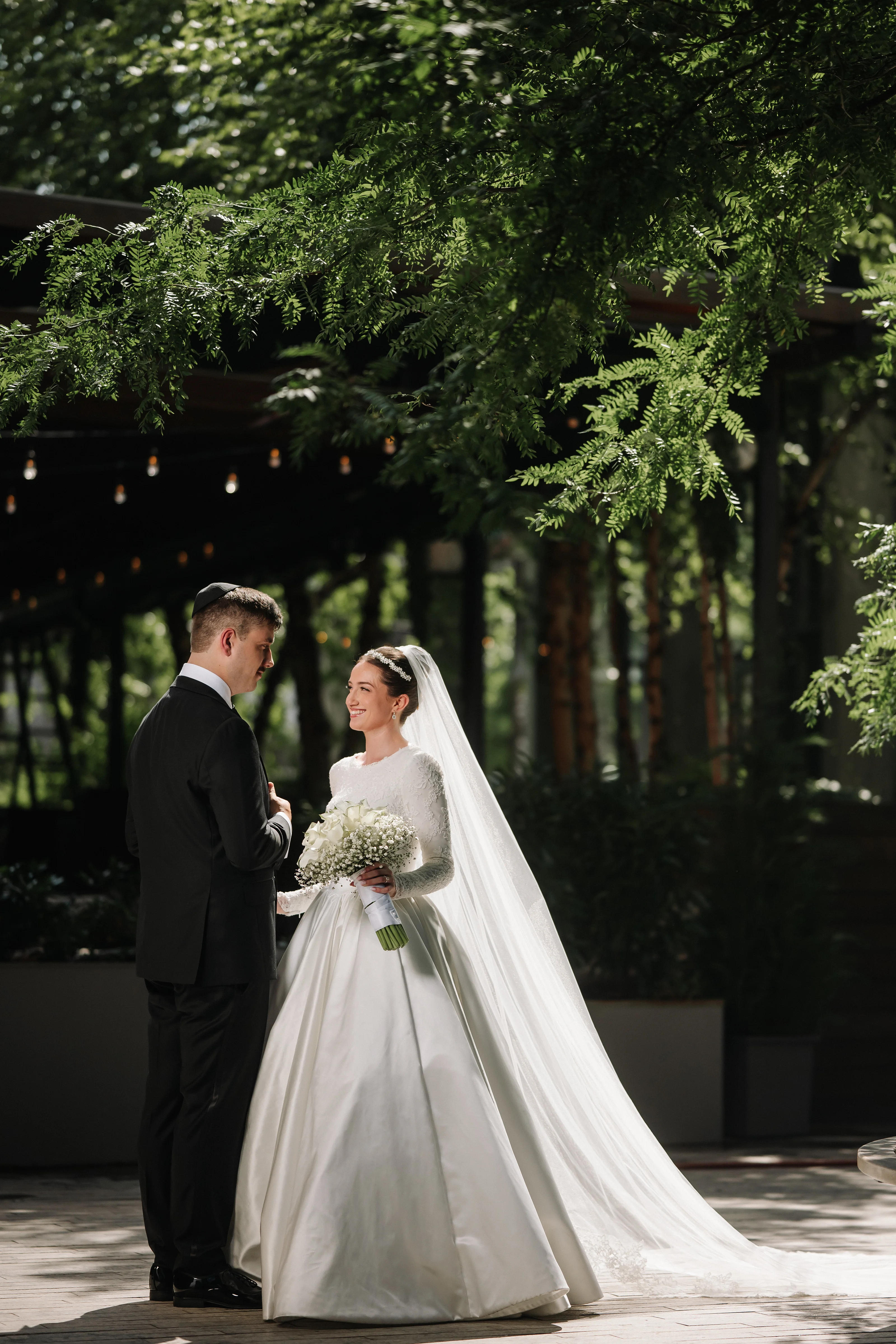 Bride and groom standing together outdoors in a garden, the bride holding a bouquet as they look at each other under leafy trees.