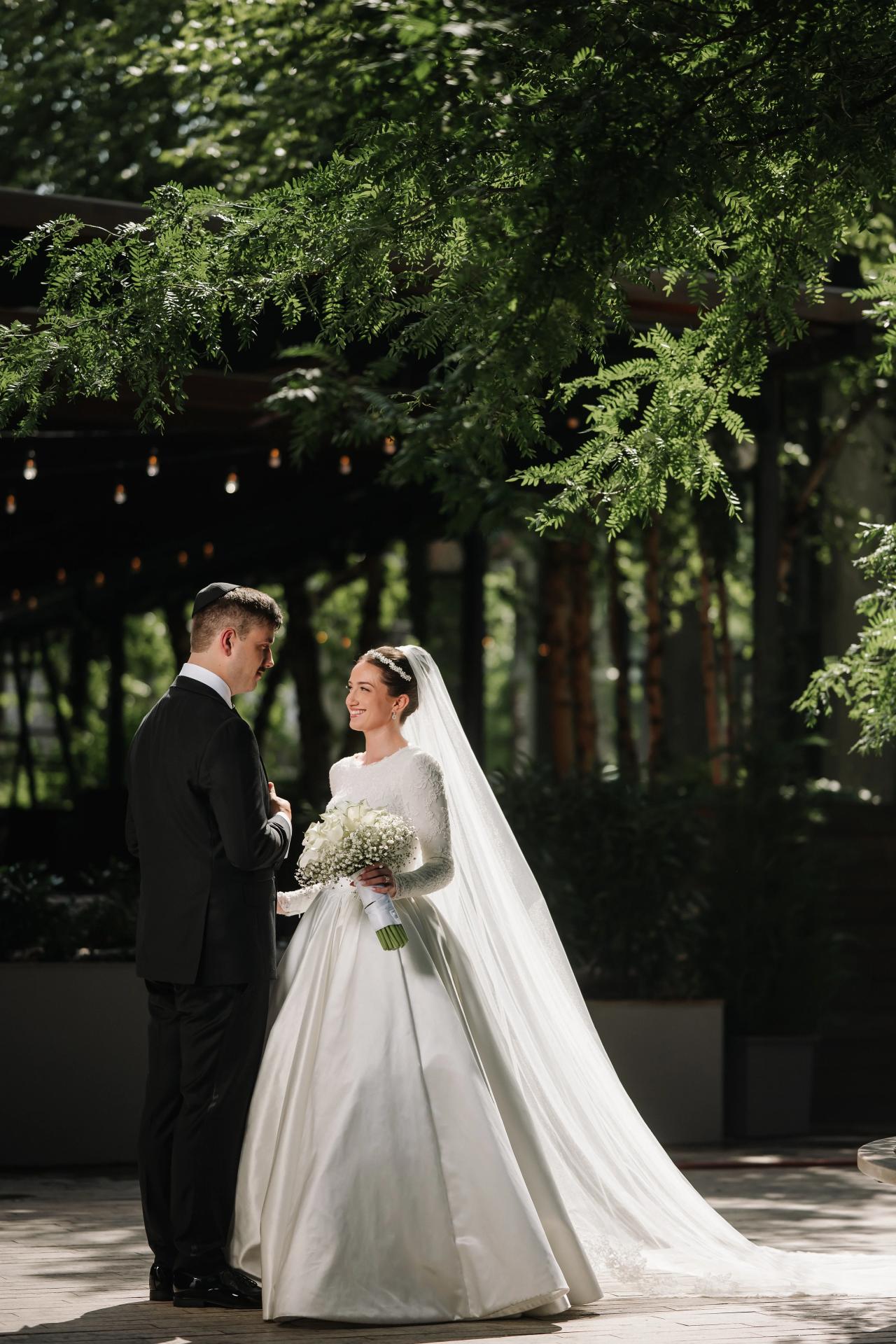 Bride and groom standing together outdoors in a garden, the bride holding a bouquet as they look at each other under leafy trees. Wedding at Ateres Chynka, Brooklyn, NY.