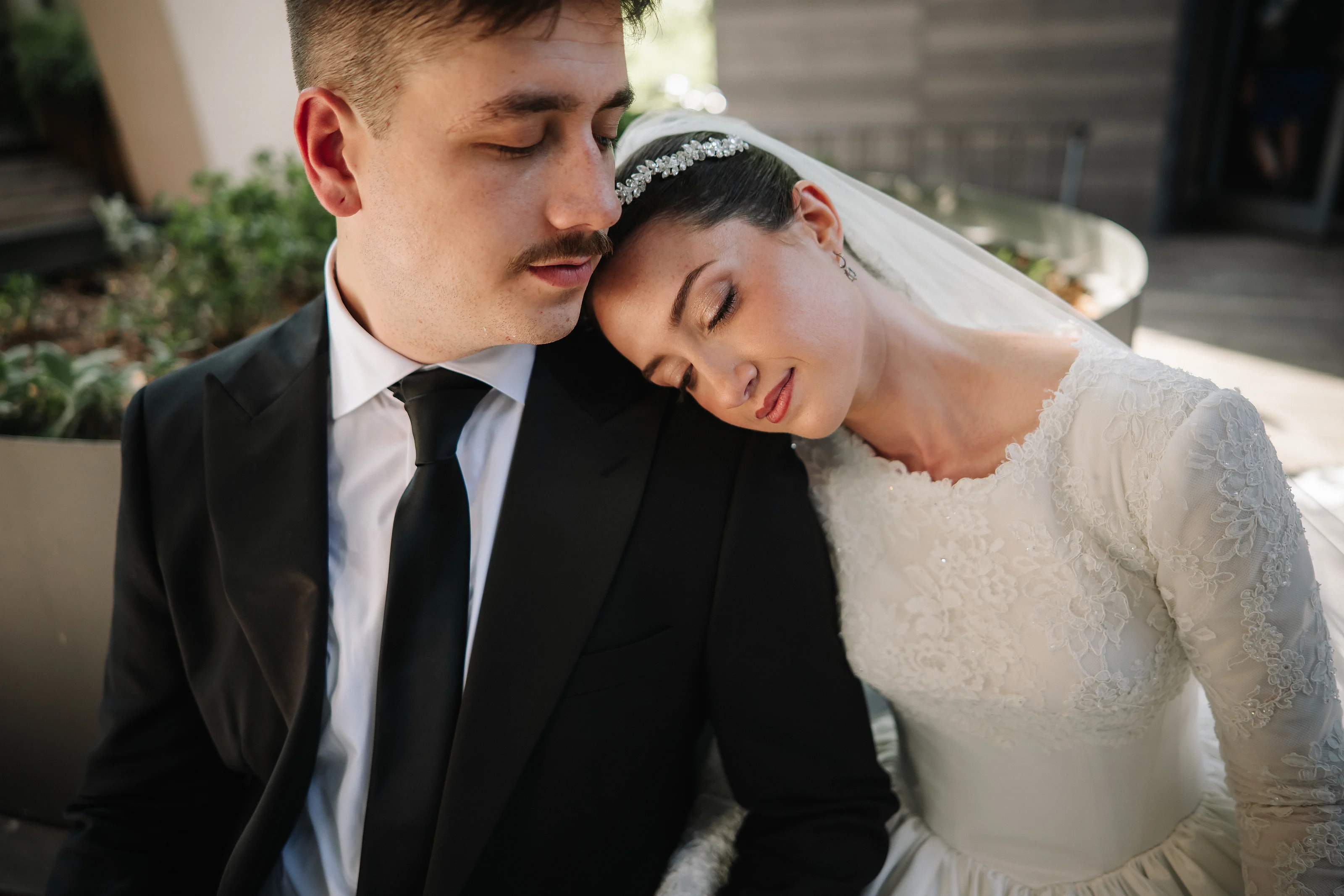 Close intimate portrait of the Jewish couple touching foreheads and leaning in, bride with veil and groom in a black suit, captured in warm soft light with a blurred garden background.