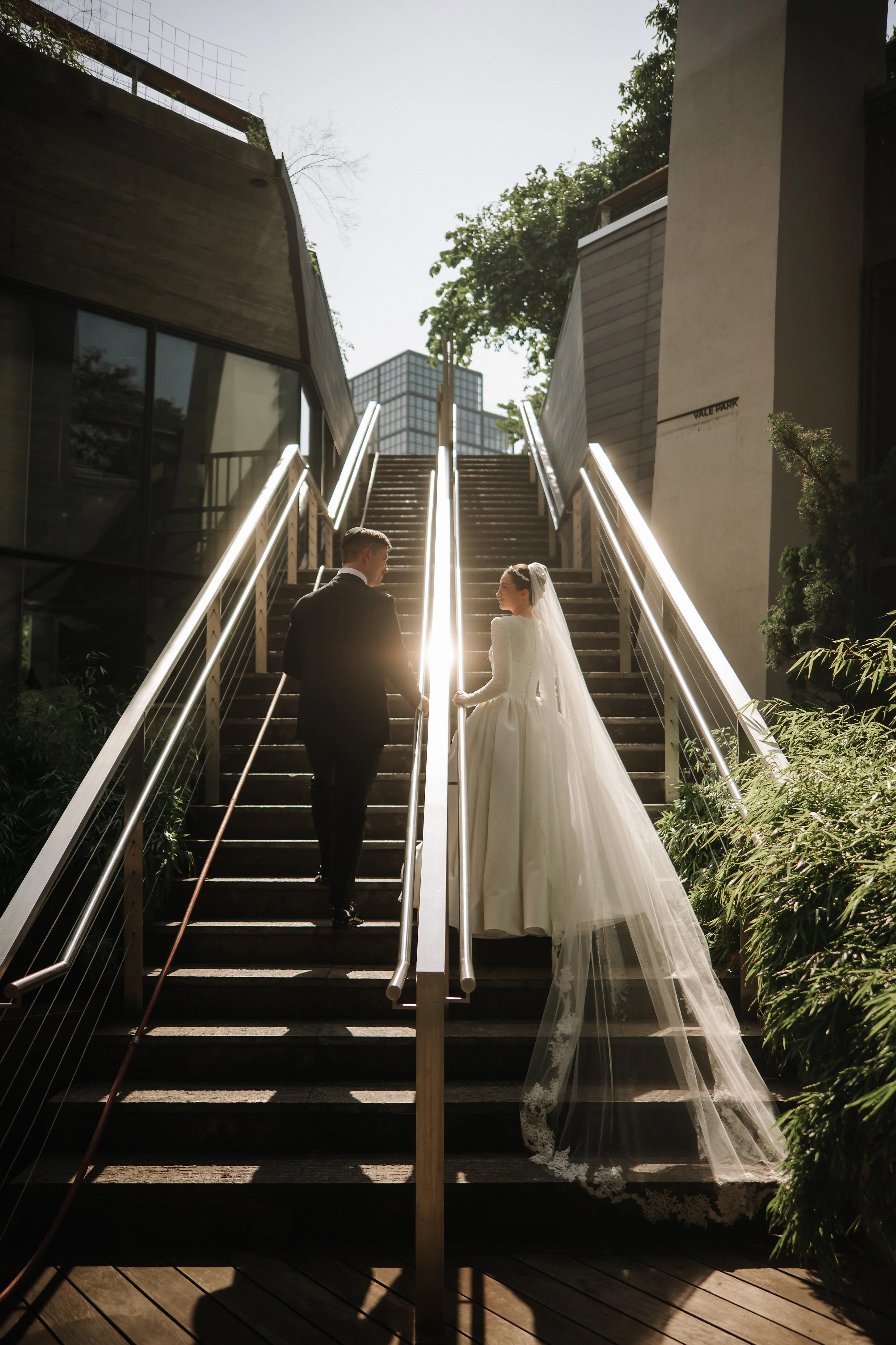 Bride in a light gown and veil walking on a sunlit staircase as the groom stands facing her, backlit by strong golden-hour light.