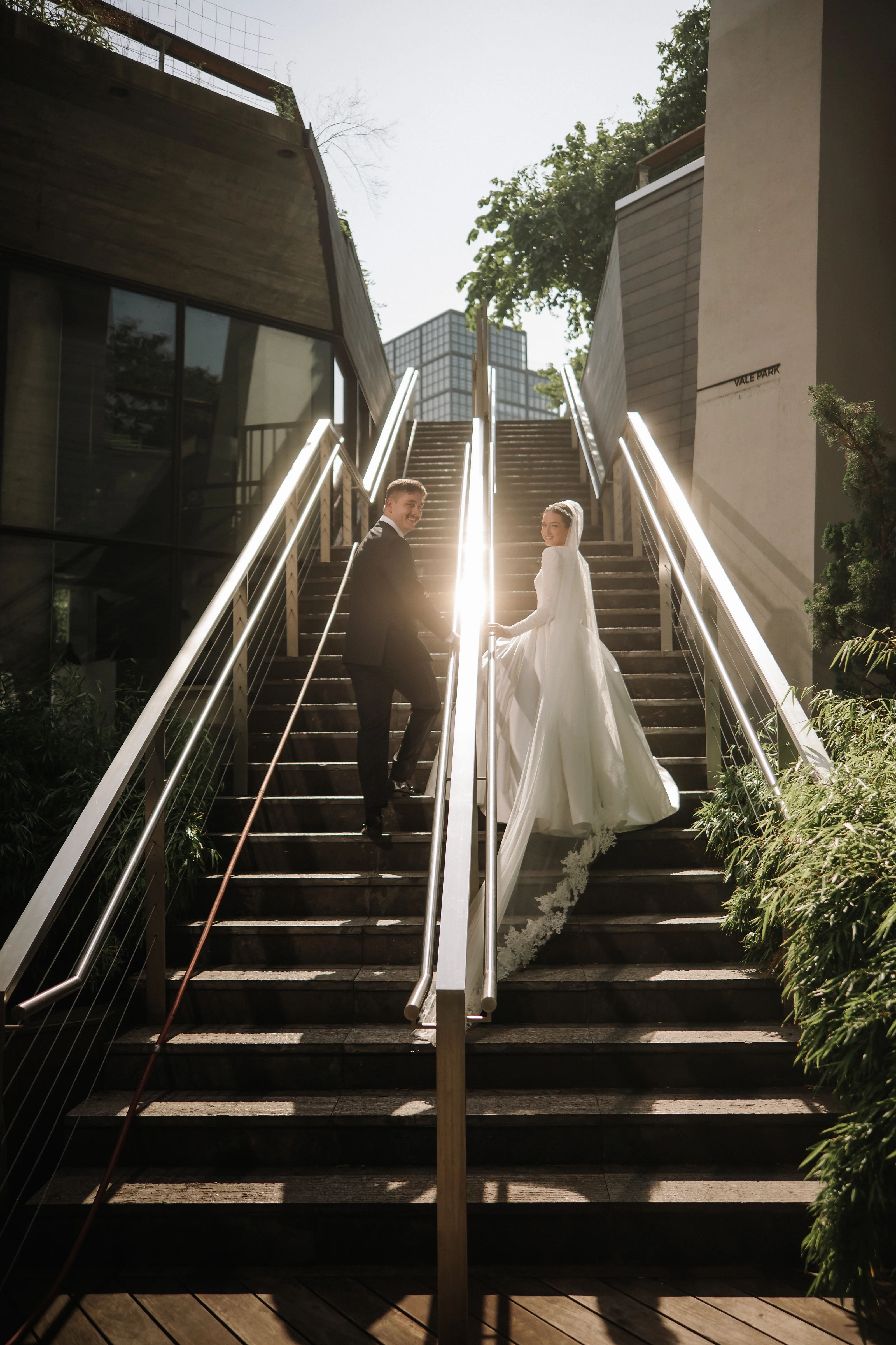 Bride and groom walking down sunlit outdoor steps during the badeken, with dramatic beams of light framing the couple.