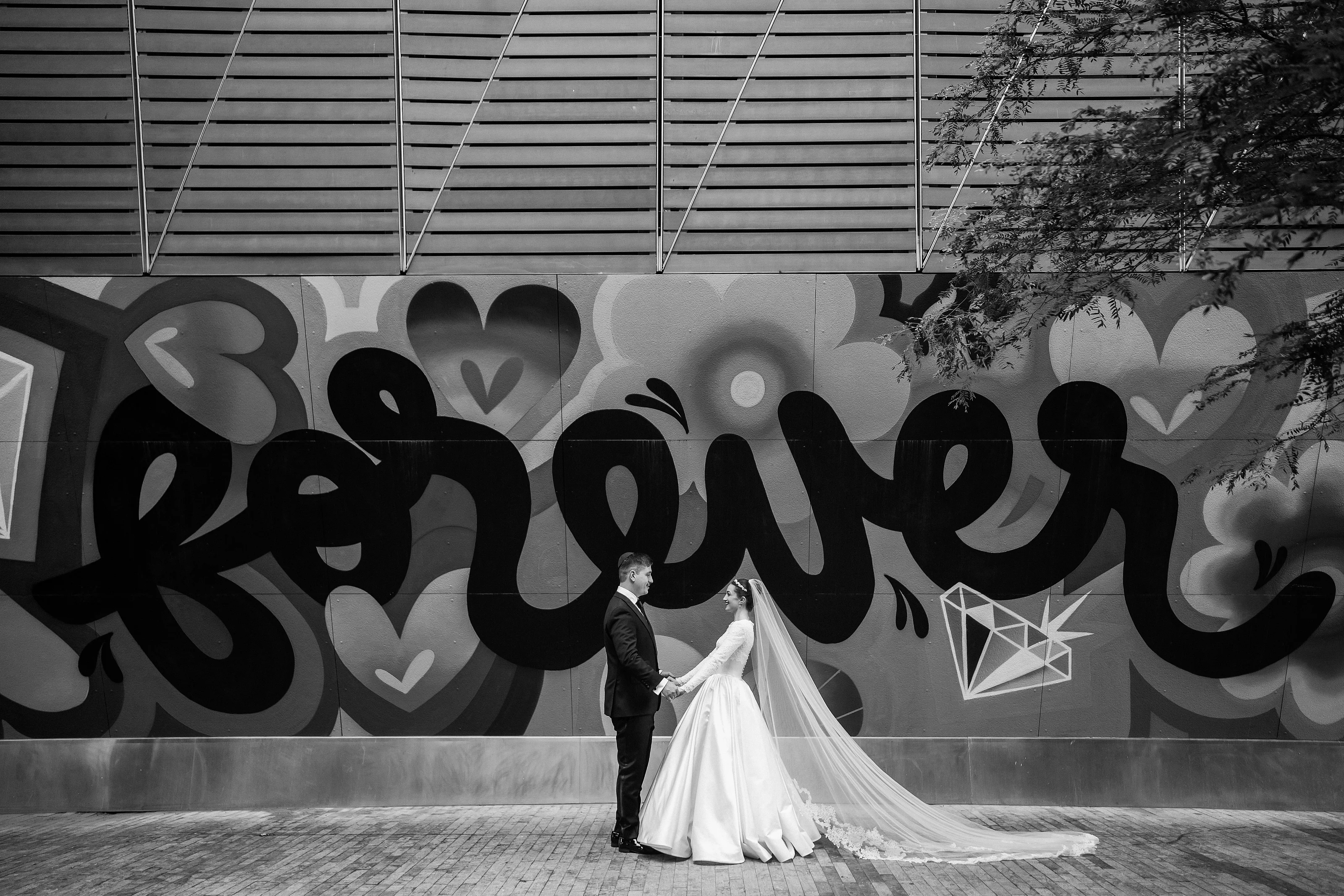 Black-and-white wedding photograph of a bride and groom standing close together with the bride’s veil flowing, posed against a large graffiti-style mural and hanging lights.