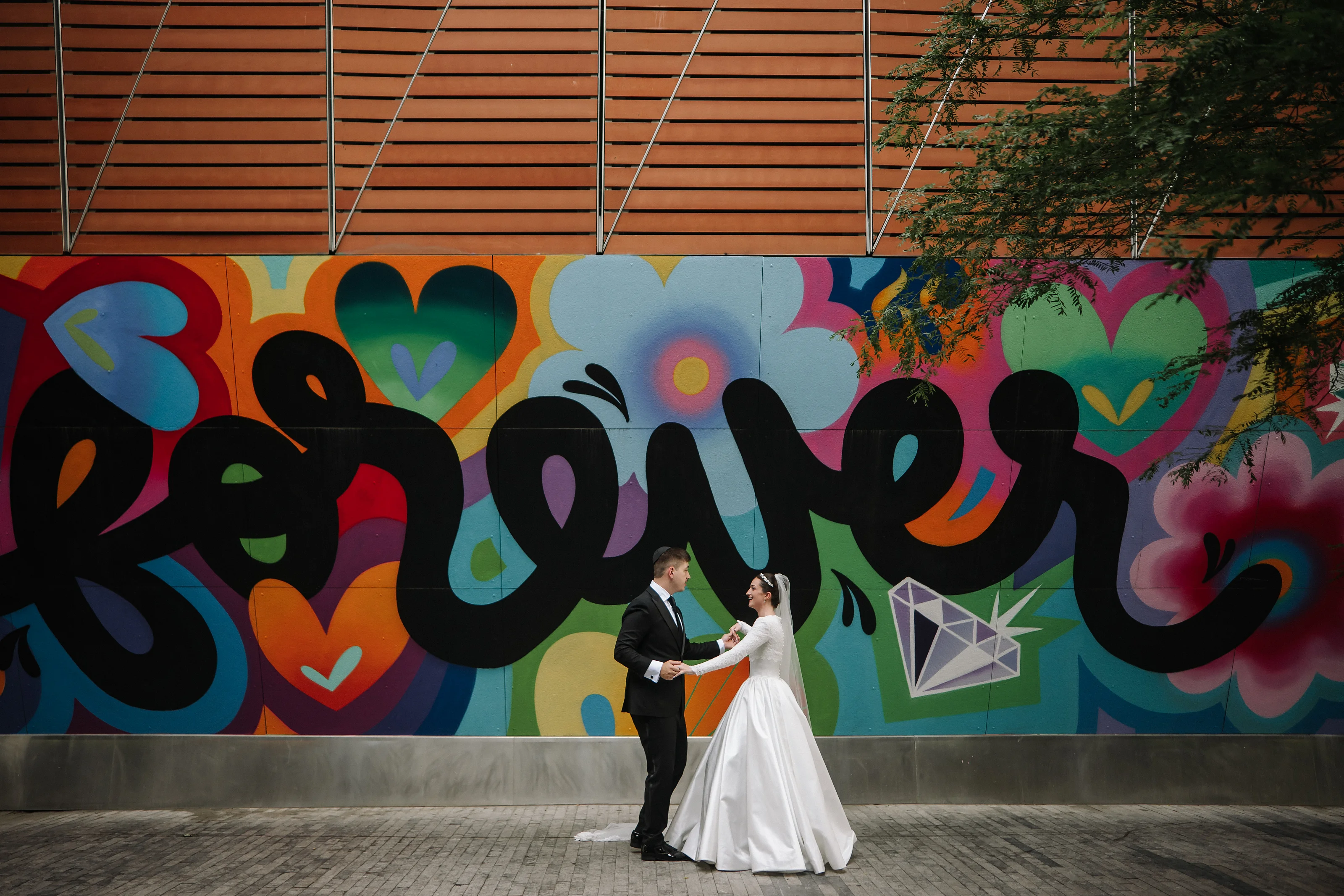 Bride and groom holding hands and walking together in front of a colorful graffiti mural backdrop.
