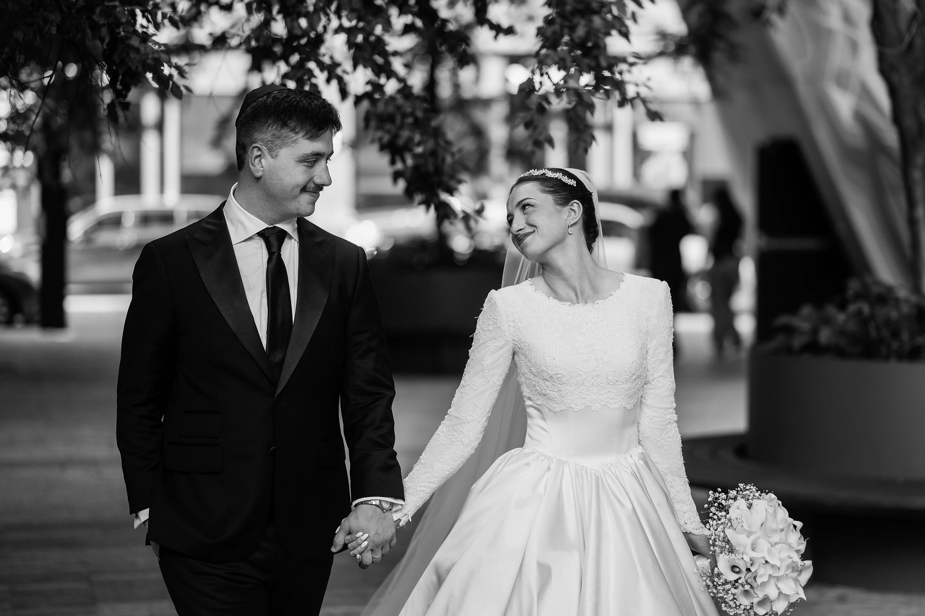 Black-and-white wedding portrait of a bride and groom holding hands and smiling as they walk under trees, with the bride holding a bouquet.