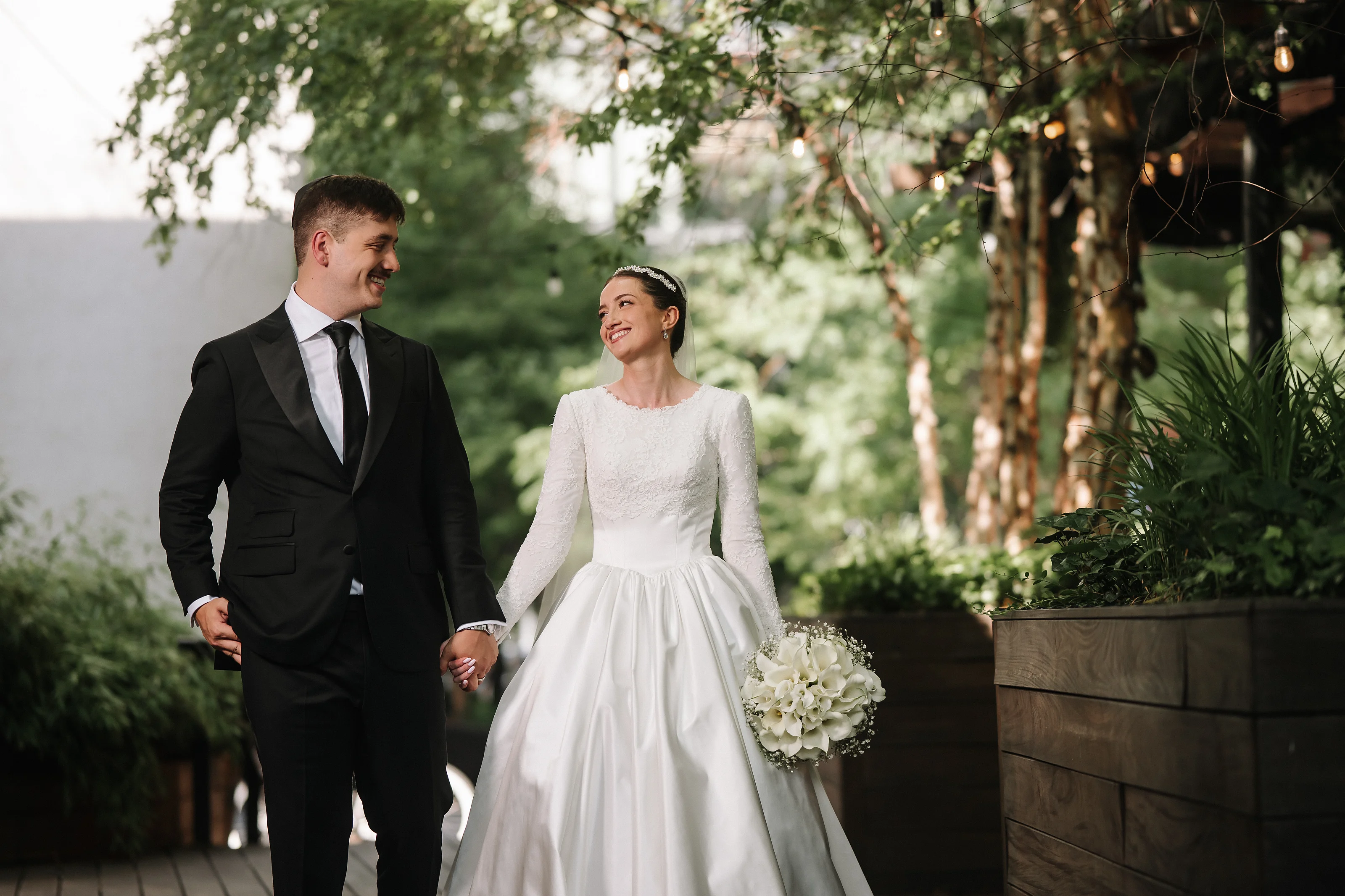 Bride and groom holding hands outdoors in a garden during golden-hour style light, smiling at each other.