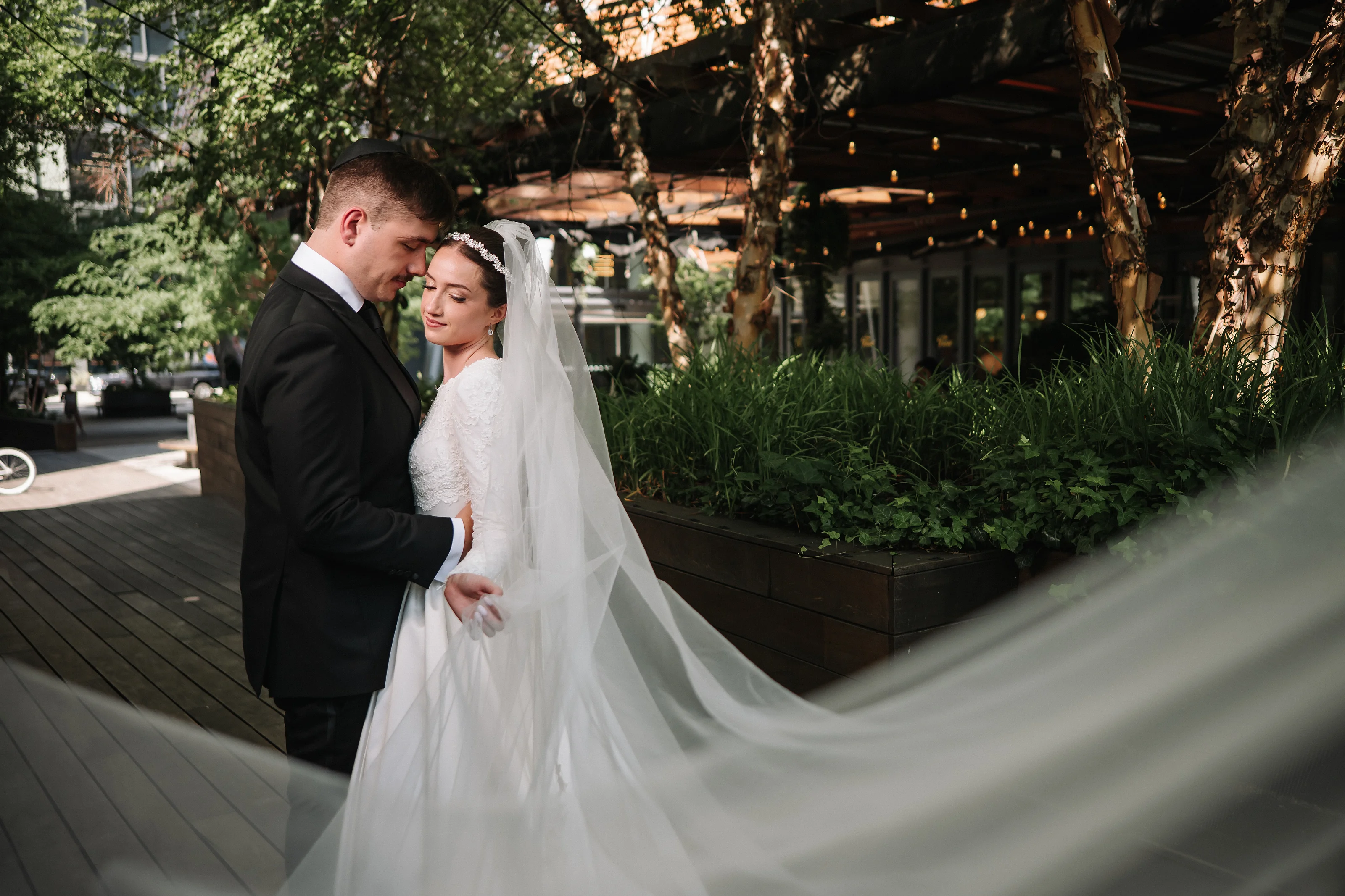 Bride in a white gown with veil lifted as the groom holds her close during badeken outdoors in warm golden-hour light.