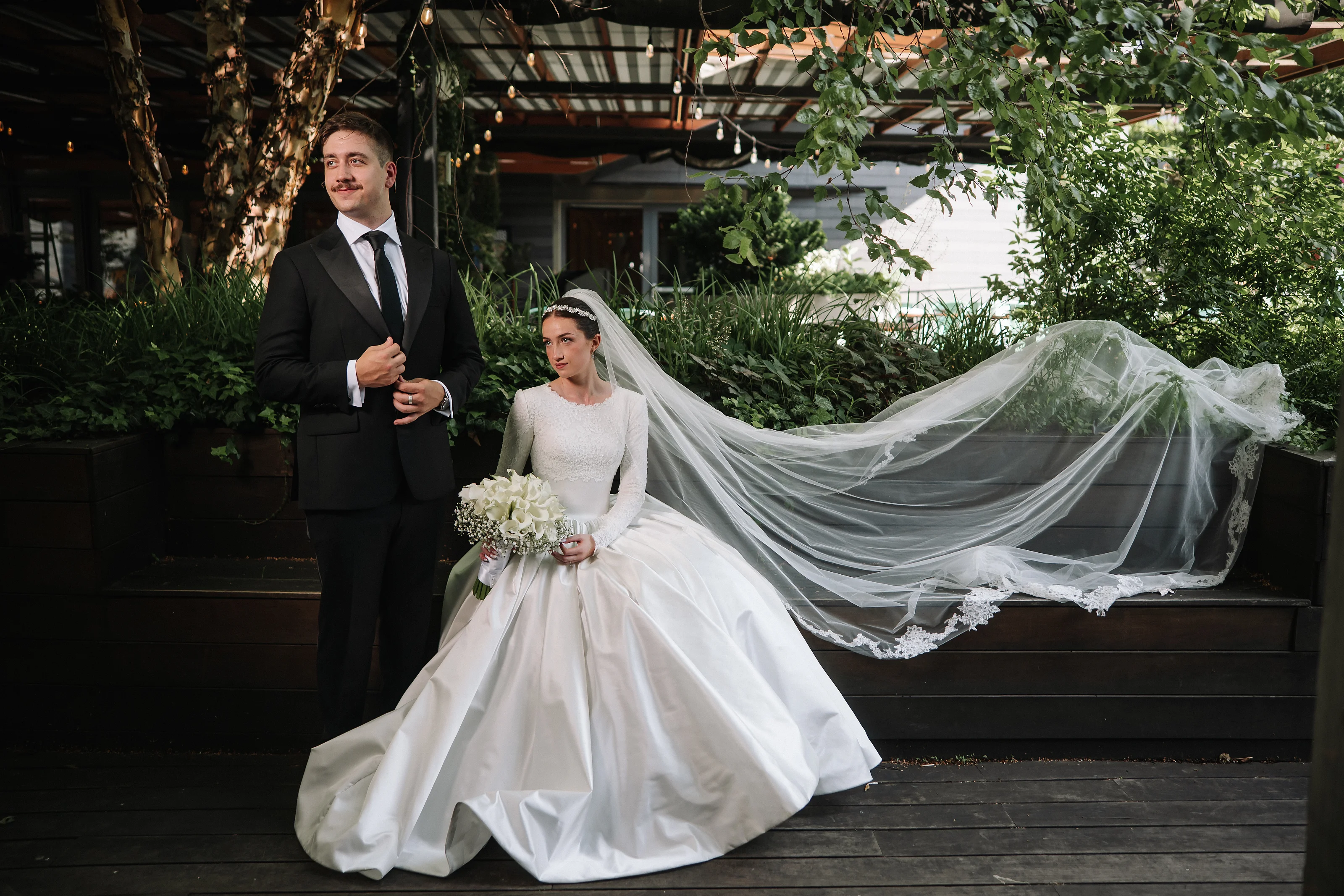 A bride and groom stand together during a wedding moment, with the bride’s veil flowing and greenery surrounding them.