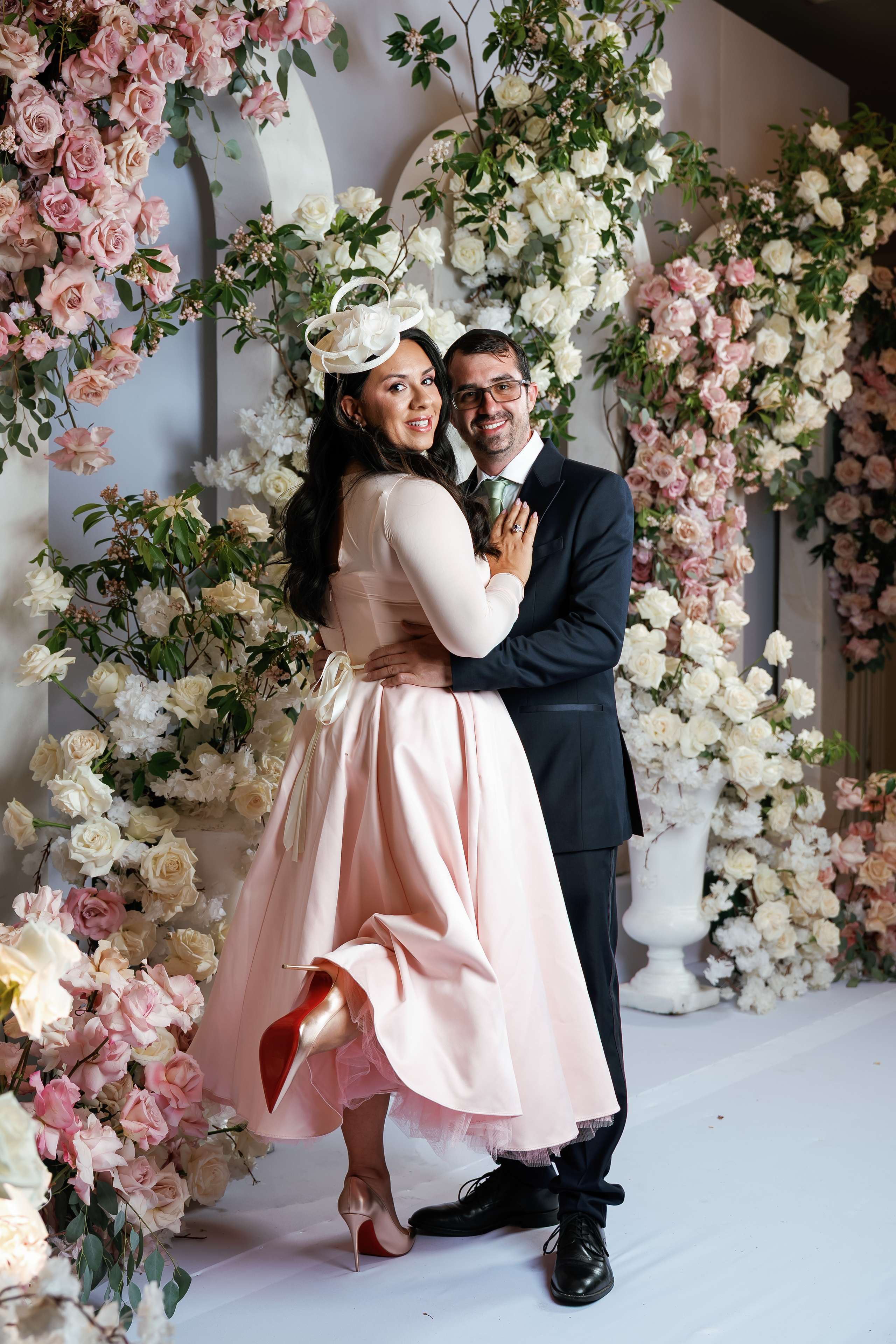 Wedding couple in a close embrace in front of an abundant pink and white floral backdrop, bride in a pale pink dress and hat, groom in a dark suit.
