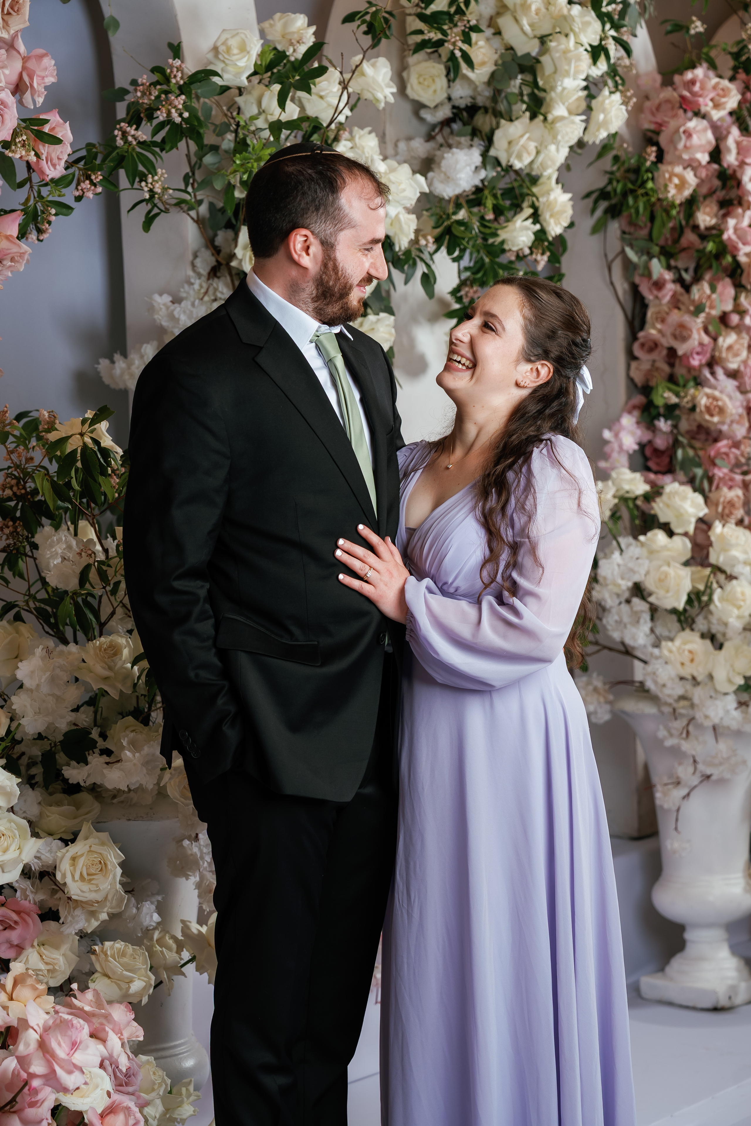 Bride and groom embracing and smiling at each other in front of a white and blush floral backdrop during a wedding ceremony.